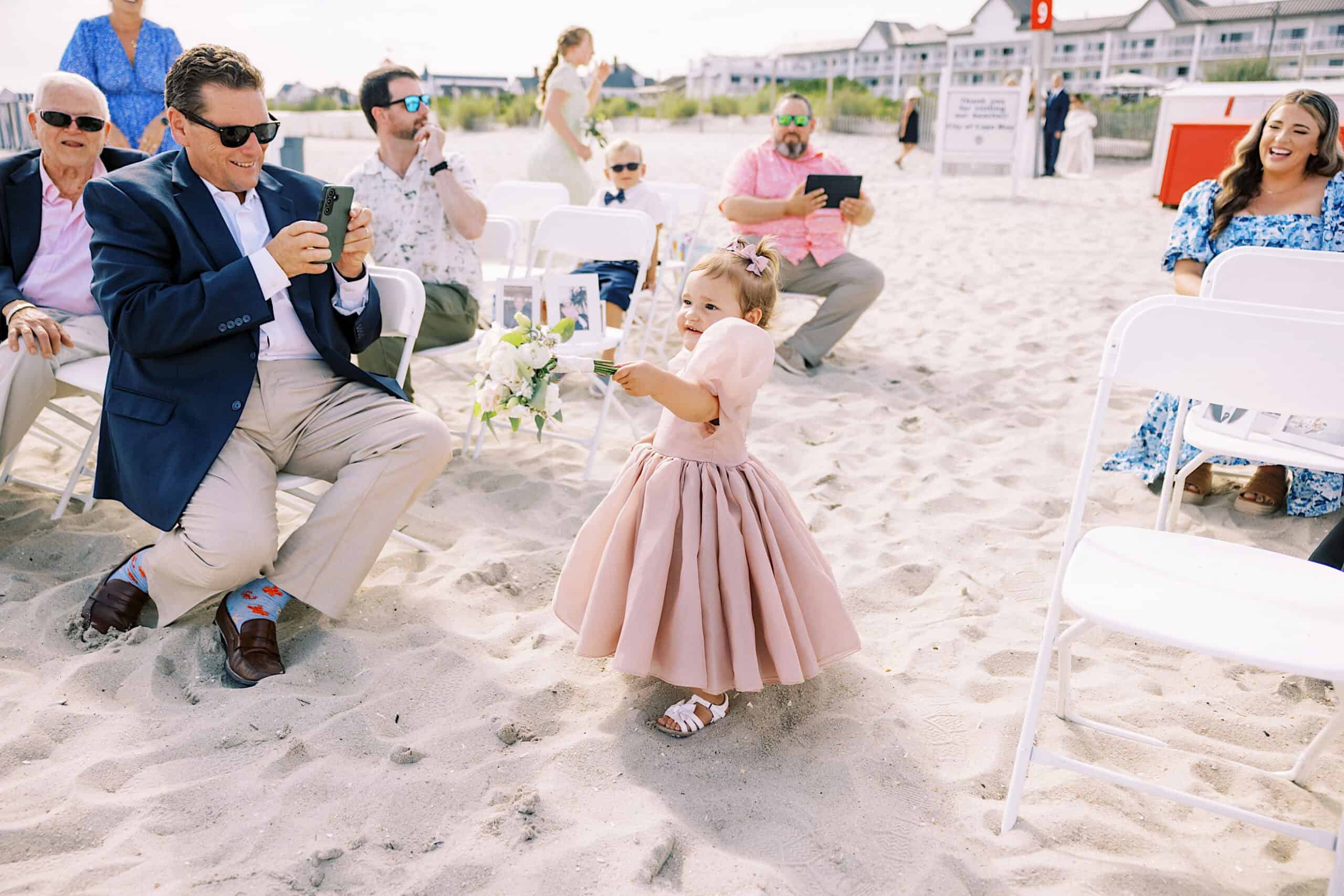 A young girl in a pink dress walks on the sand between rows of white chairs, while adults seated nearby watch and take photos at a beautiful Montreal Beach Resort Wedding.