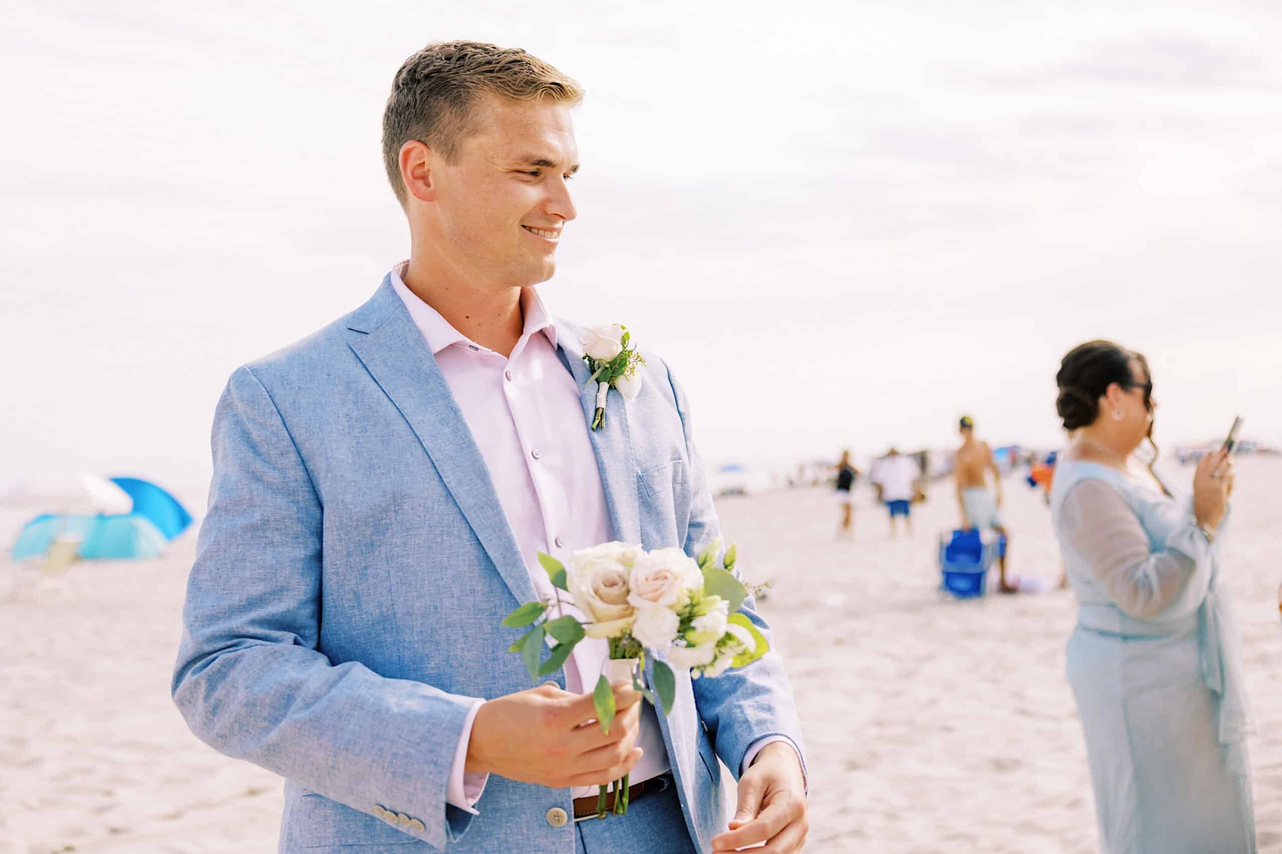 A man in a light blue suit holding a small bouquet of white roses stands on a sandy beach at a Montreal Beach Resort Wedding; people and beach umbrellas are visible in the background.