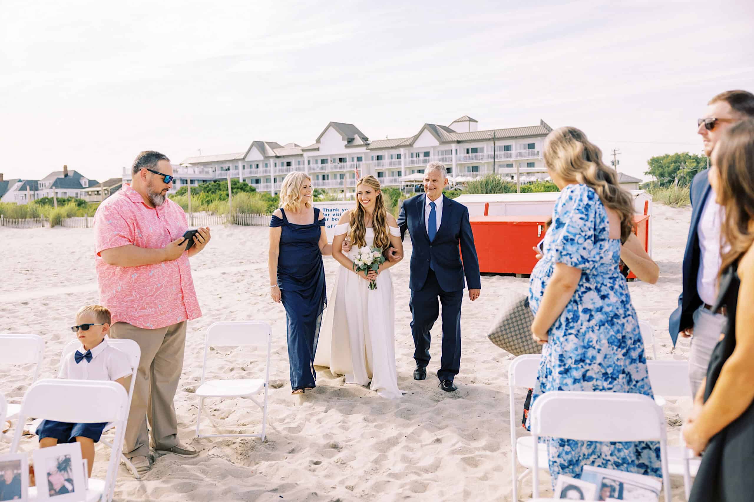 A bride walks down the aisle on a sandy beach at a Montreal Beach Resort Wedding, accompanied by two adults, while guests watch and take photos. Beach resort buildings and a red dumpster are in the background.