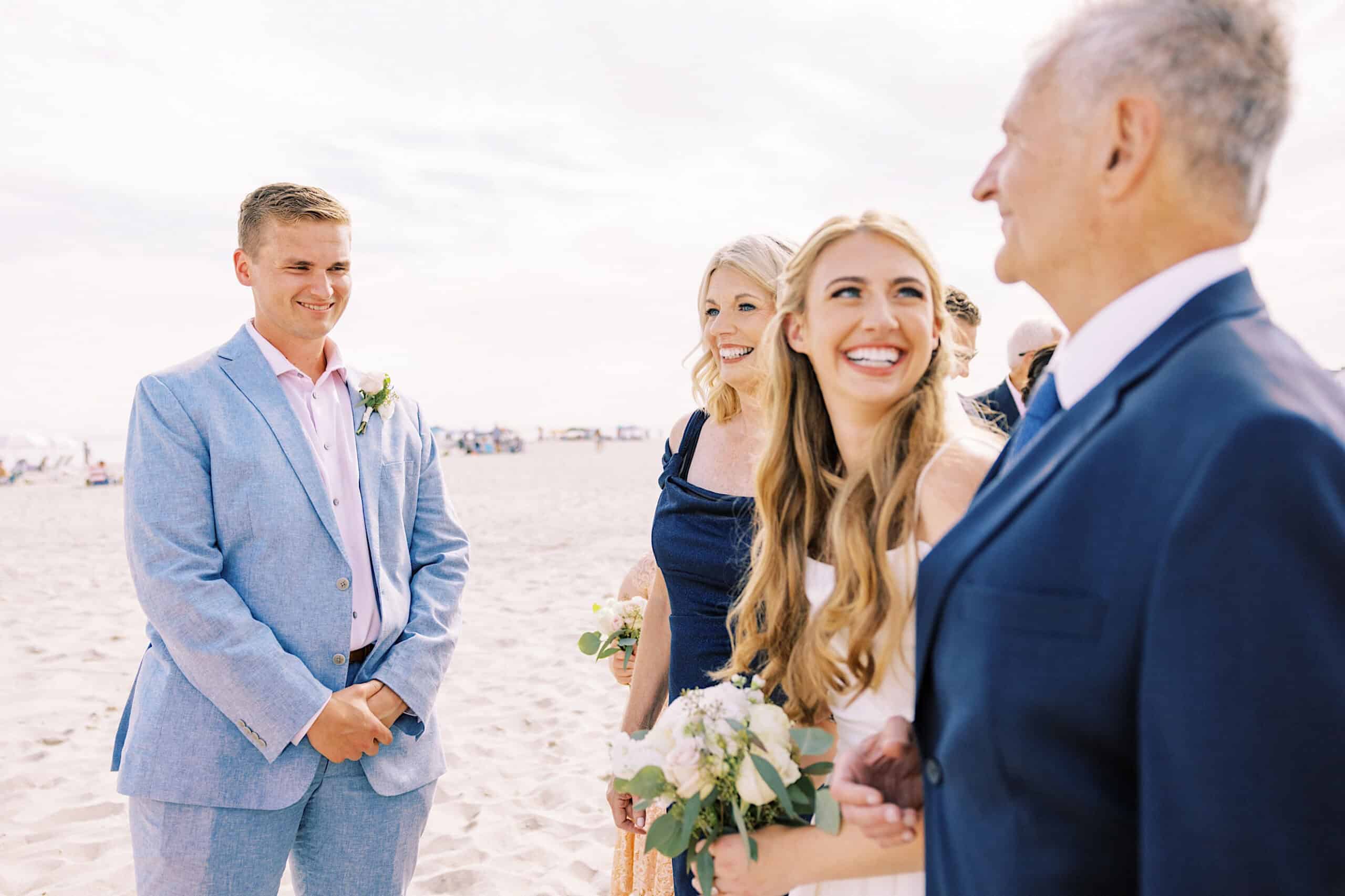 A bride holding a bouquet smiles at an older man, while a groom in a light blue suit and a woman in a navy dress stand beside them on the sandy shore during their Montreal Beach Resort Wedding.