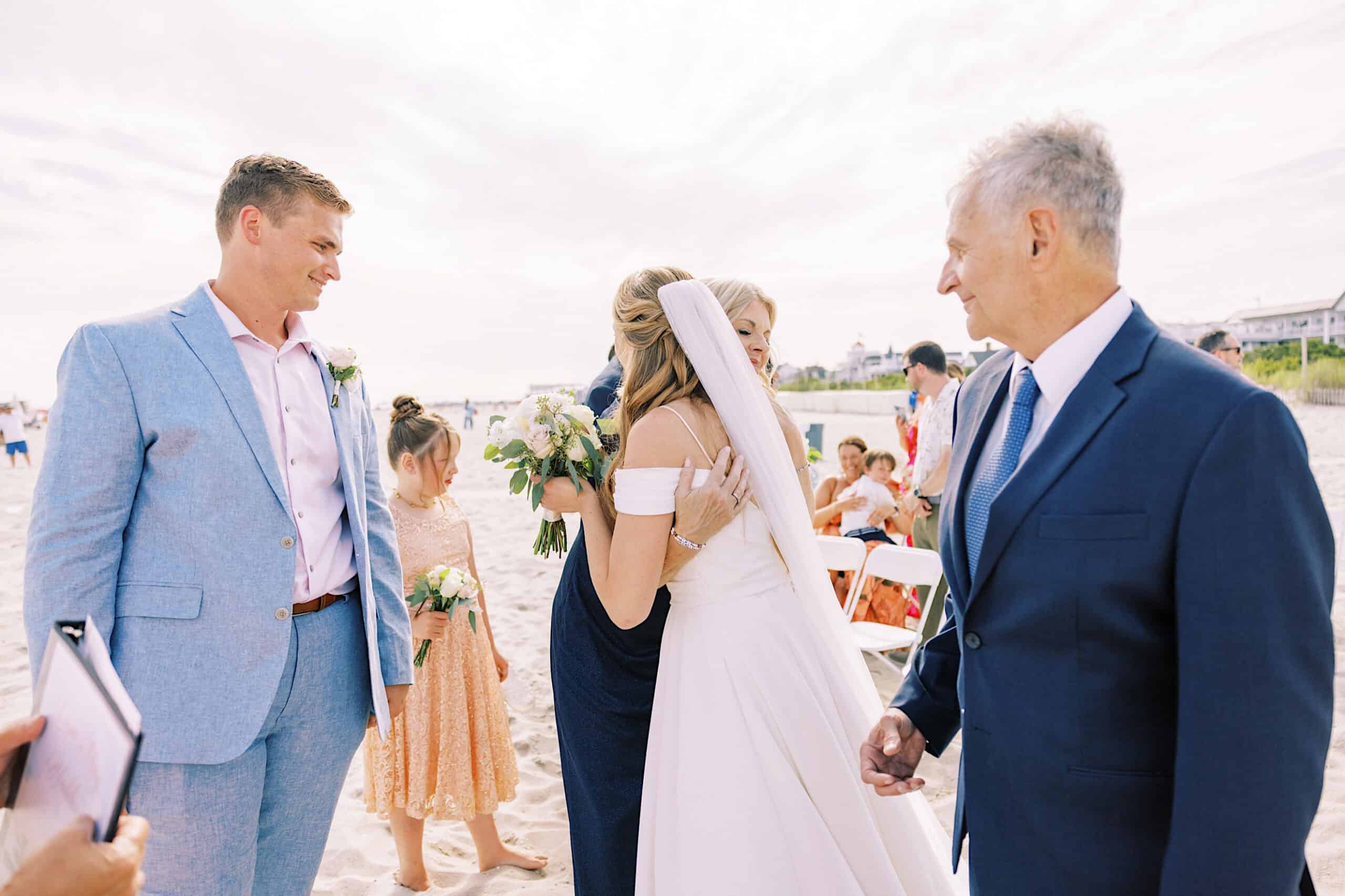 A bride in a white dress hugs a woman on the sandy shore during a Montreal Beach Resort Wedding, as guests in formal attire look on. Nearby, a man in a blue suit stands while the flower girl holds her bouquet.