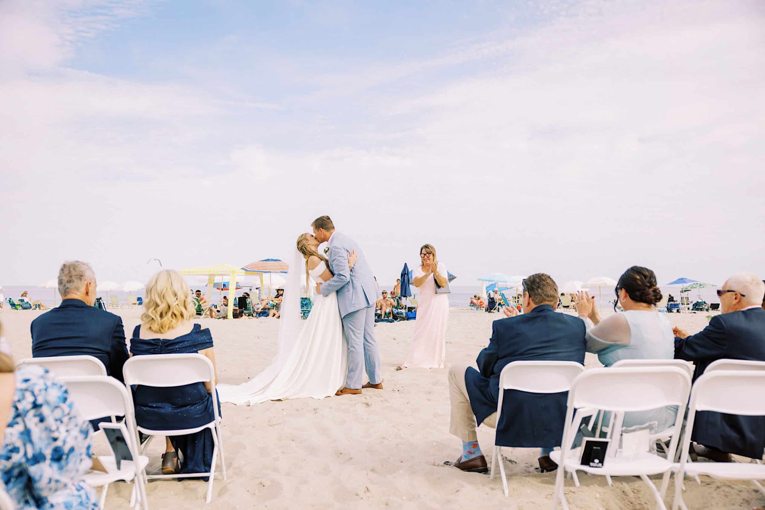 A bride and groom kiss during a Montreal Beach Resort Wedding ceremony, with guests seated on white chairs in the sand and beachgoers in the background.