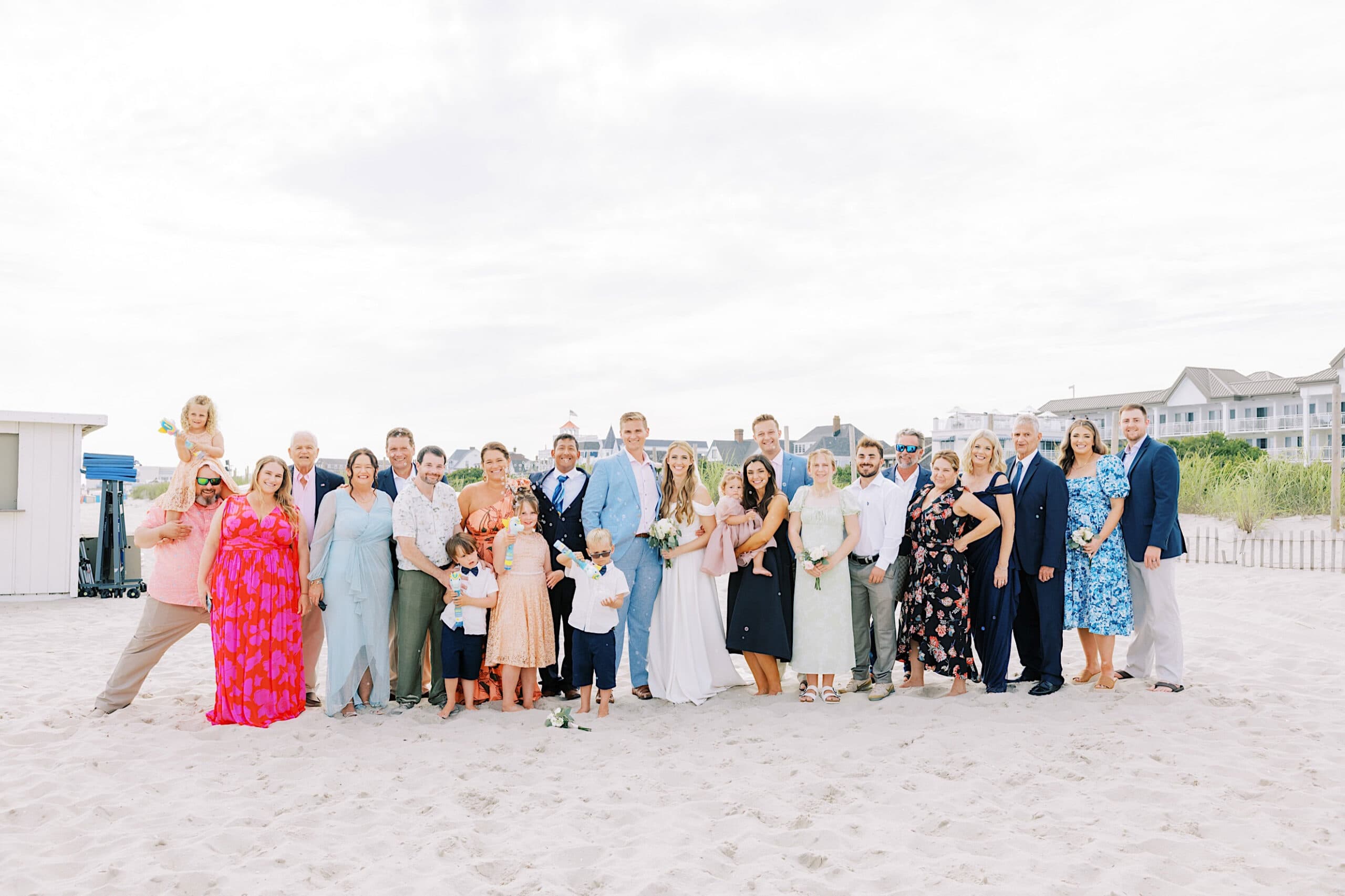 A large group of adults and children pose together on a sandy beach at a Montreal Beach Resort wedding, dressed in formal and semi-formal attire, with buildings and greenery in the background.