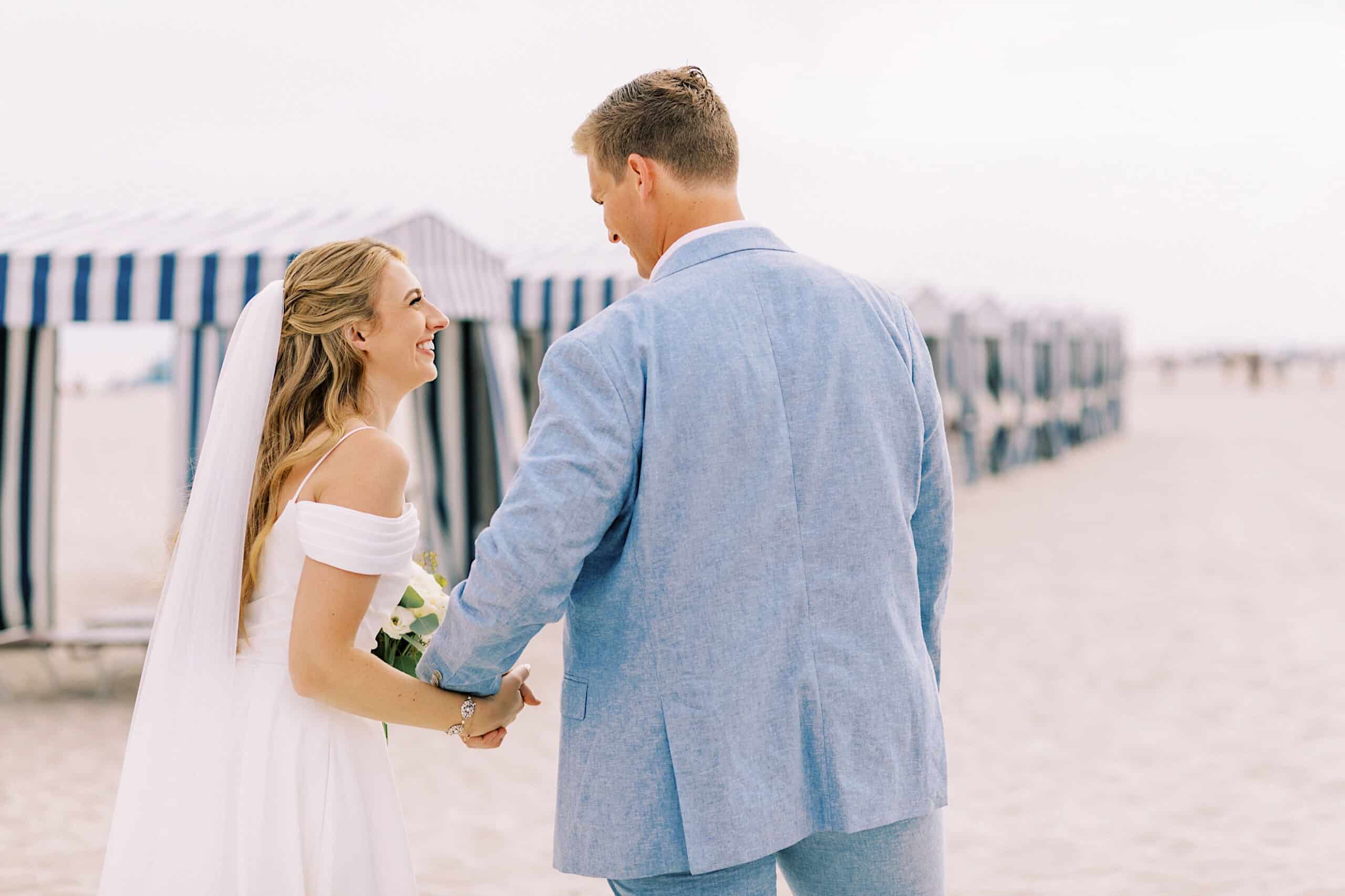 A bride and groom hold hands and smile at each other while standing on a sandy beach with striped cabanas in the background during their Montreal Beach Resort Wedding.