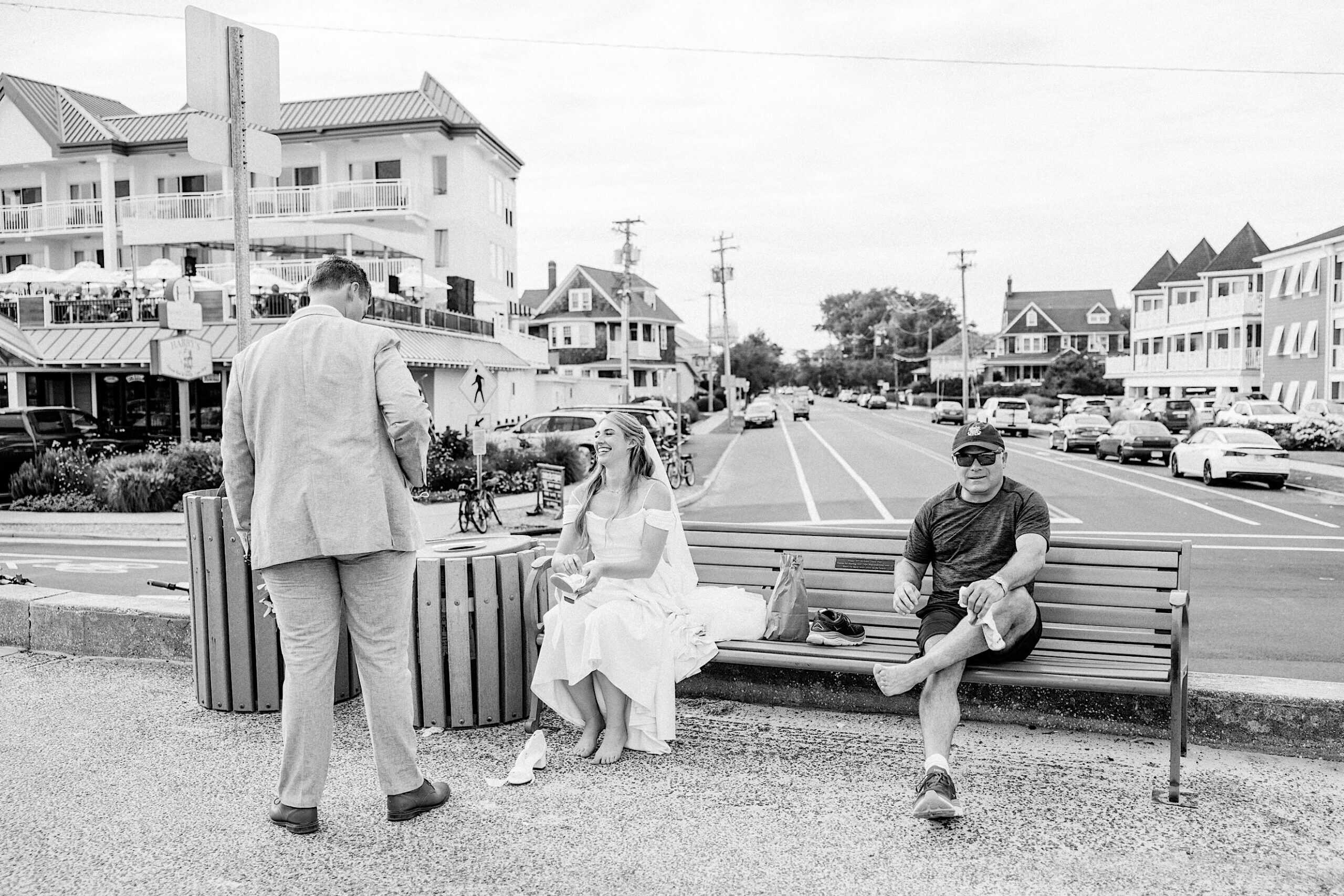 A bride in a dress and veil sits on a bench next to a man in casual clothes while a man in a suit stands nearby, capturing the relaxed spirit of a Montreal Beach Resort Wedding on a street lined with buildings and parked cars.