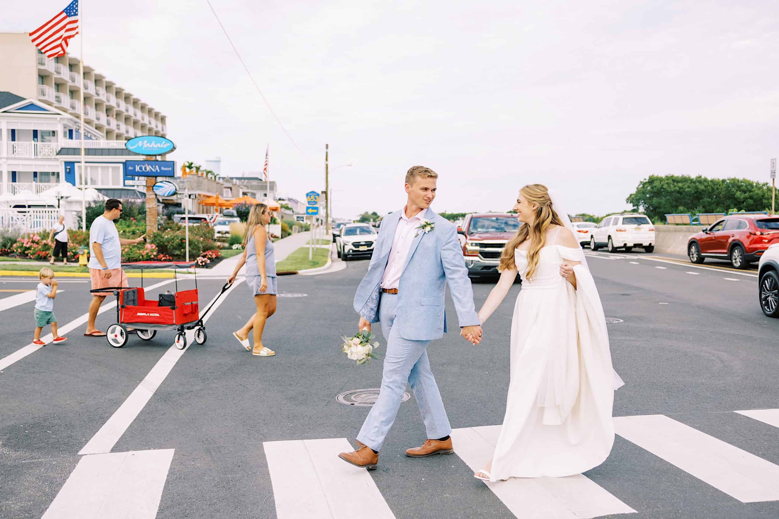 A bride and groom hold hands while crossing a street at a crosswalk during their Montreal Beach Resort wedding; other pedestrians and cars are visible in the background.