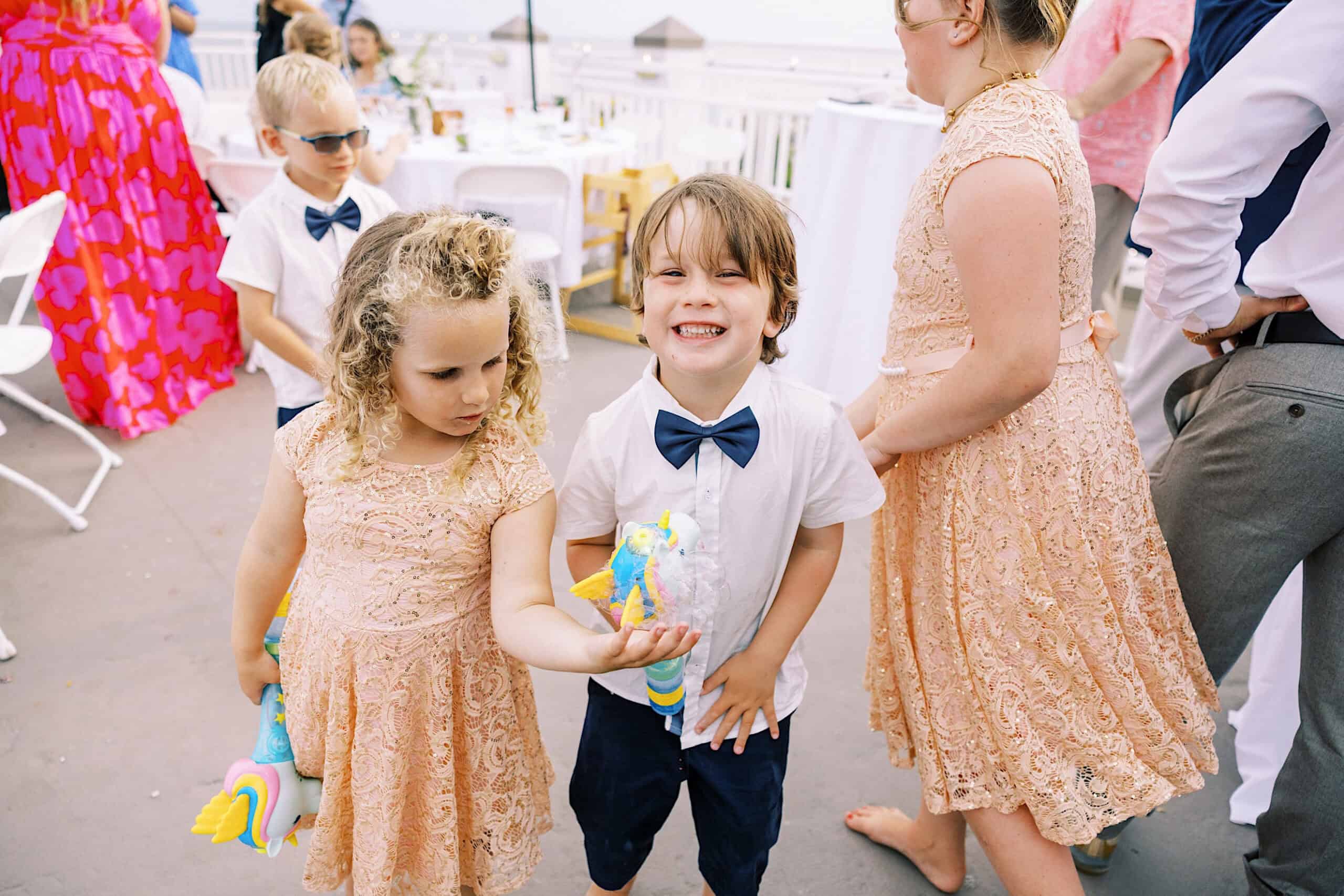 Children in light-colored formal attire stand together outdoors at a Montreal Beach Resort wedding; one girl holds a toy while a boy in a bow tie smiles at the camera. Other guests mingle in the background.