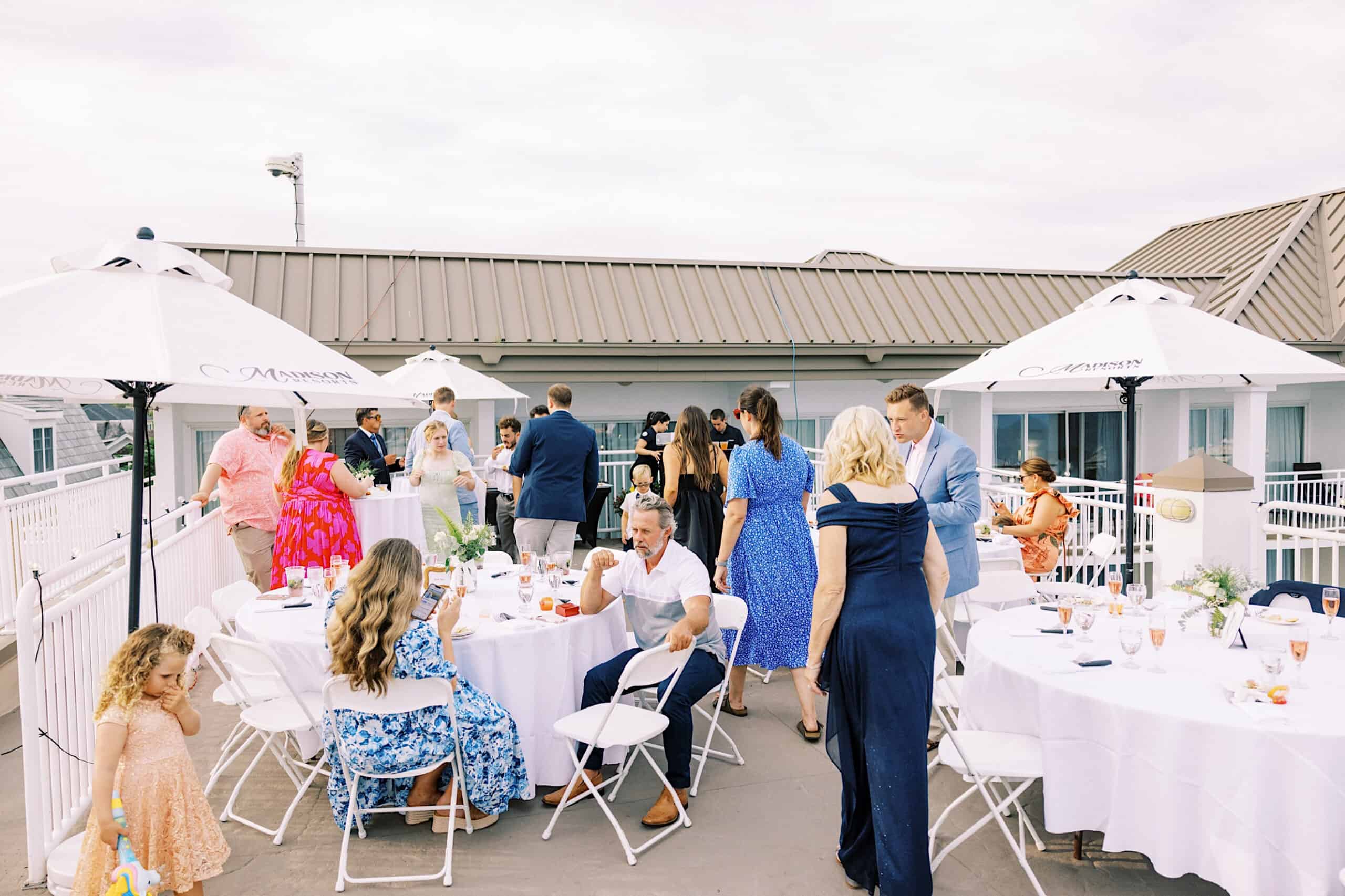 Guests mingle around white tables with umbrellas on a rooftop patio, enjoying lively conversation and dining during a Montreal Beach Resort Wedding daytime event.