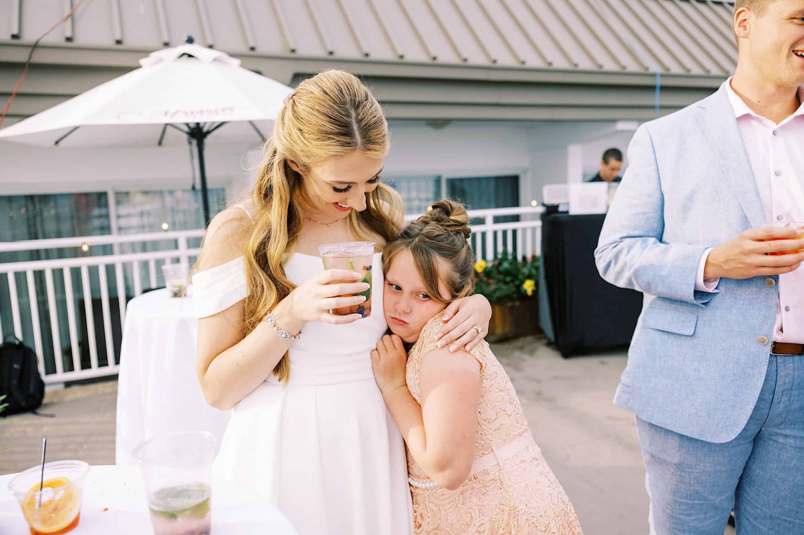 At a Montreal Beach Resort wedding, a woman in a white dress hugs a young girl in a peach dress at an outdoor event. The girl looks pensive while the woman smiles, with other people and tables in the background.
