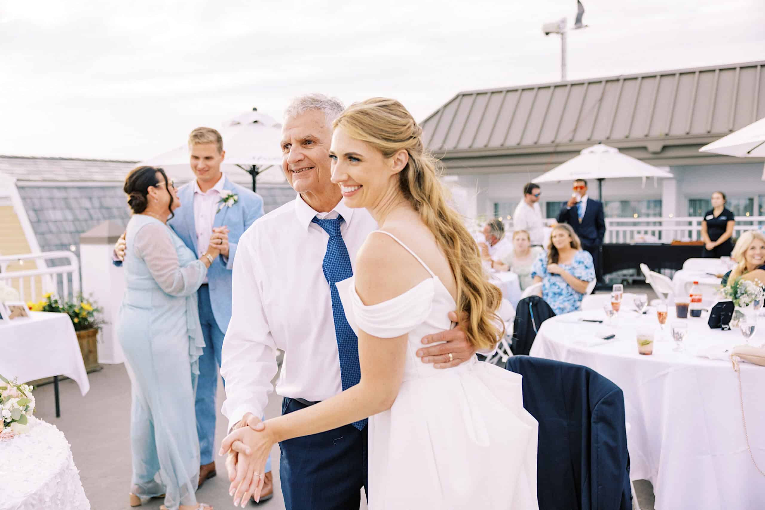 A bride in a white dress dances with an older man at an outdoor Montreal Beach Resort Wedding, with guests seated and standing in the background.