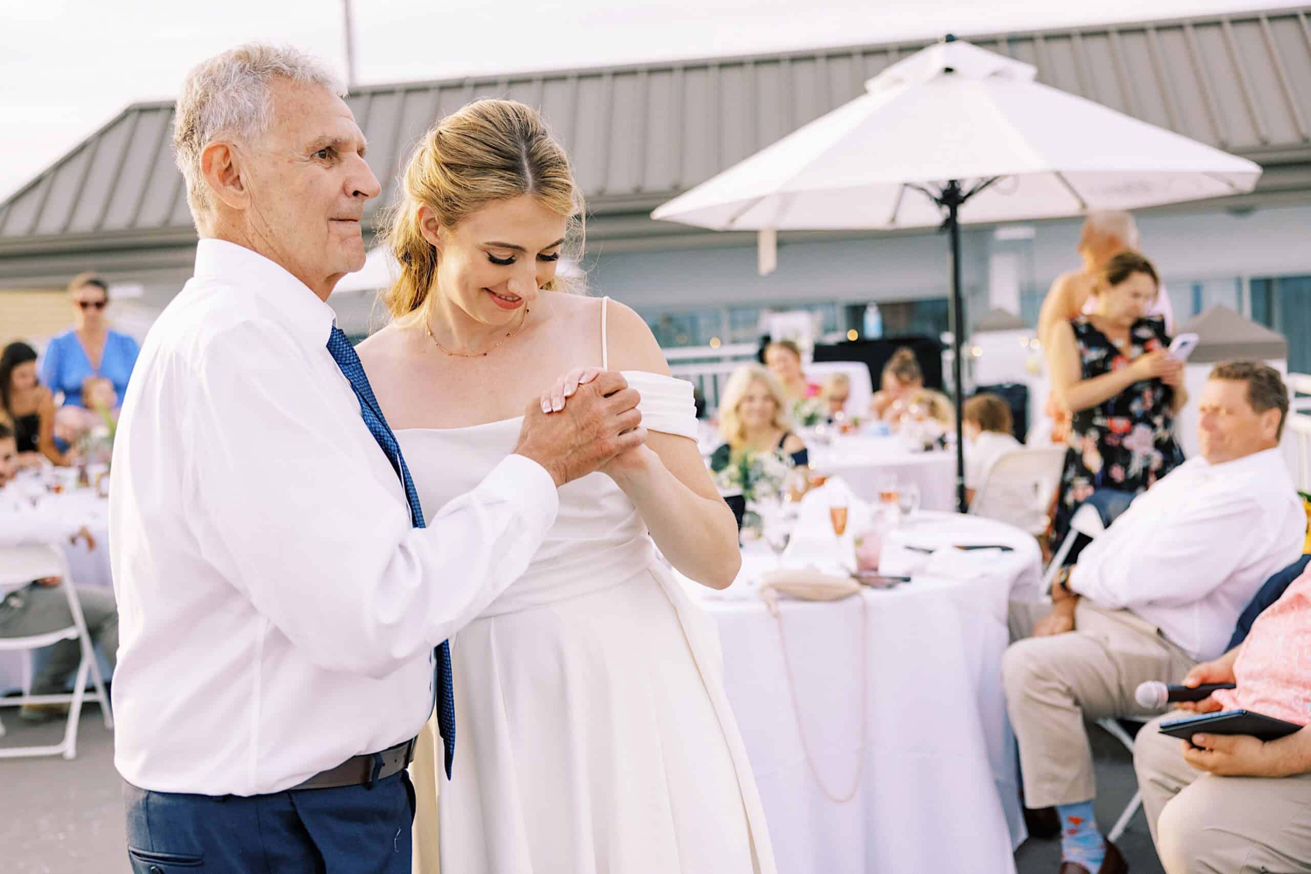 At a Montreal Beach Resort Wedding, a bride in a white dress dances with an older man in a white shirt and tie at an outdoor reception, while guests watch from round tables.