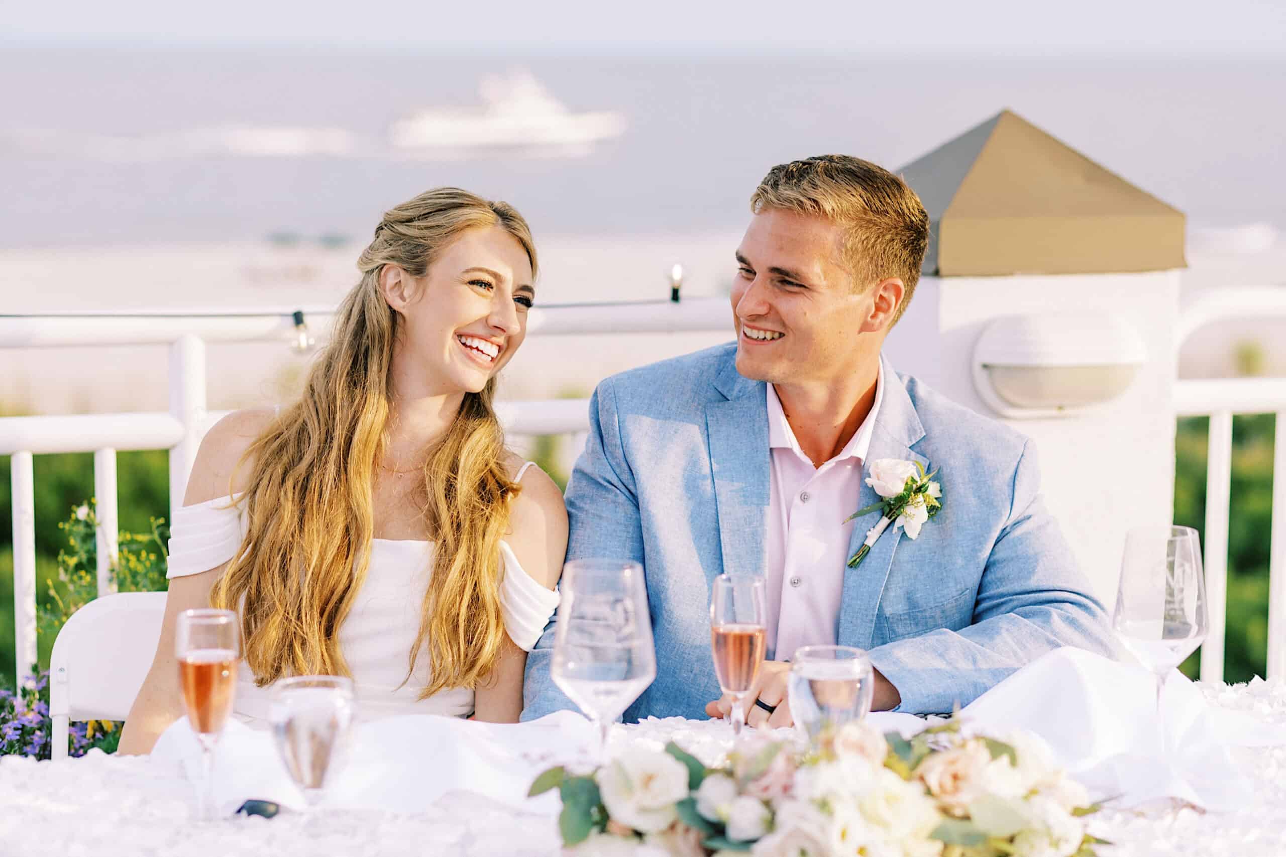 A woman in a white dress and a man in a light blue suit sit at a decorated table outdoors, smiling at each other, with drinks and flowers in front of them and water in the background, capturing the charm of a Montreal Beach Resort Wedding.