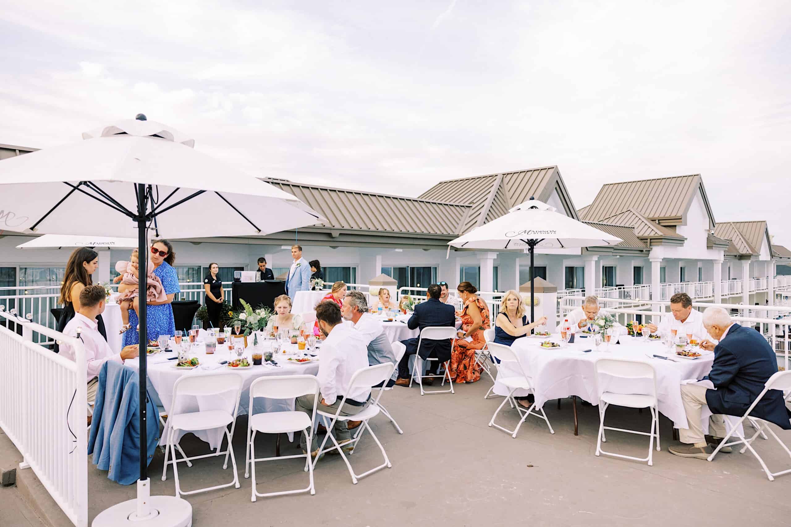 Guests gather at round tables with white tablecloths under umbrellas on a rooftop patio, enjoying food and conversation during a daytime Montreal Beach Resort Wedding.