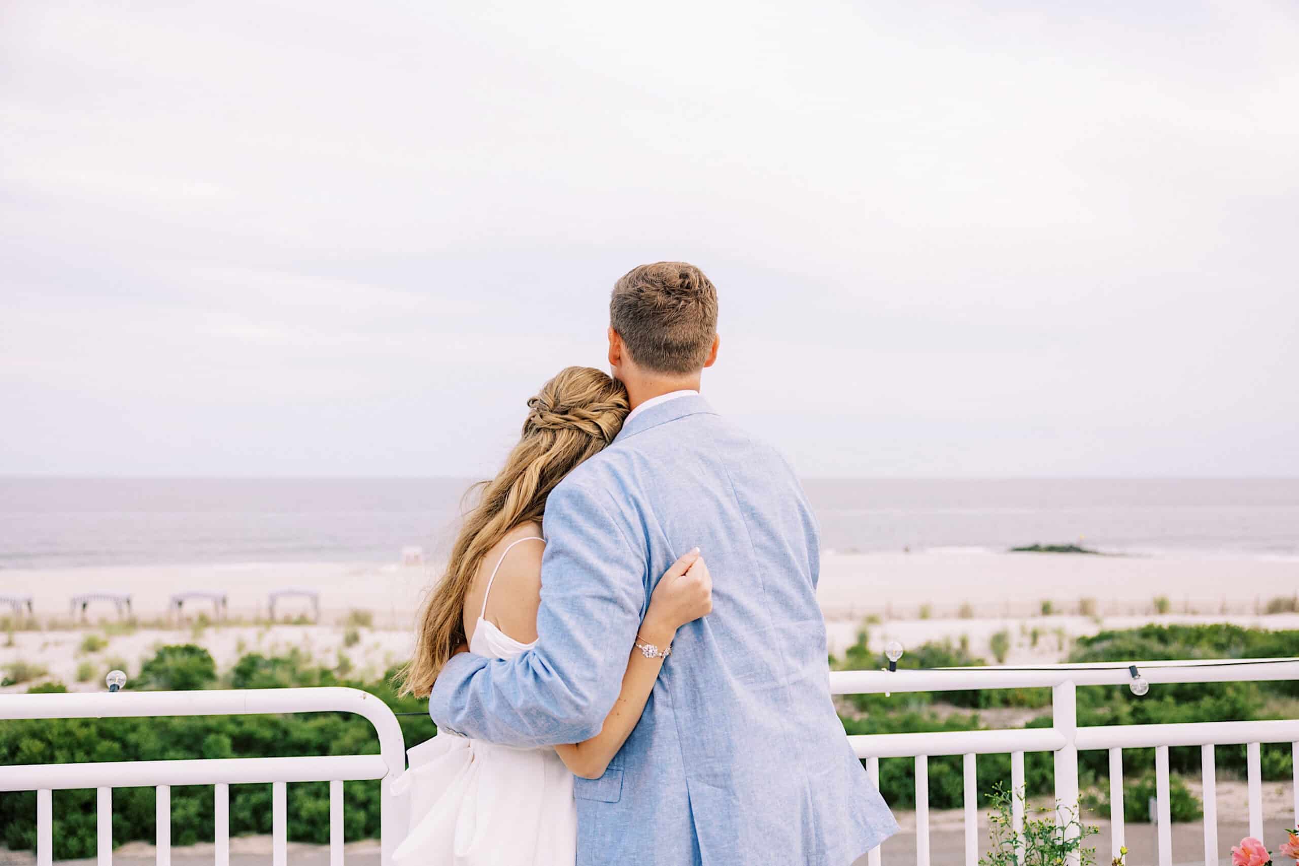 A couple stands on a balcony at Montreal Beach Resort, embracing and looking out at a sandy beach and the ocean under a cloudy sky, capturing the magic of their wedding day.
