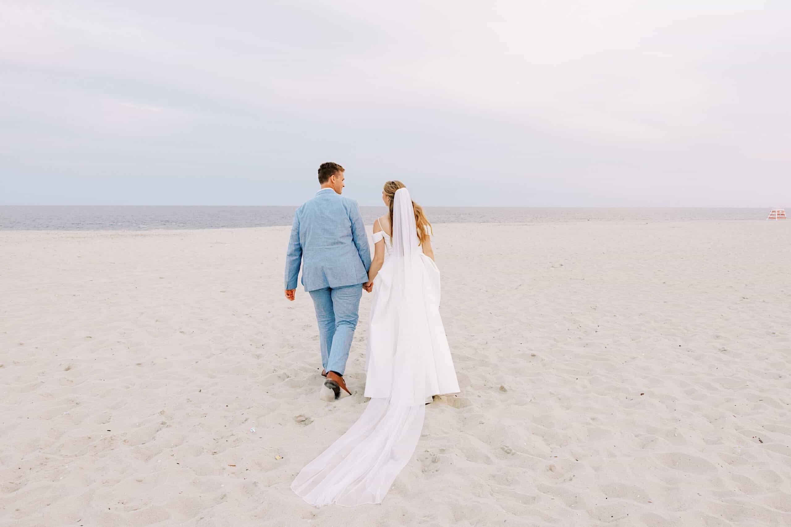 A couple dressed in wedding attire walks hand in hand on a sandy beach toward the ocean under a cloudy sky, enjoying their Montreal Beach Resort Wedding.