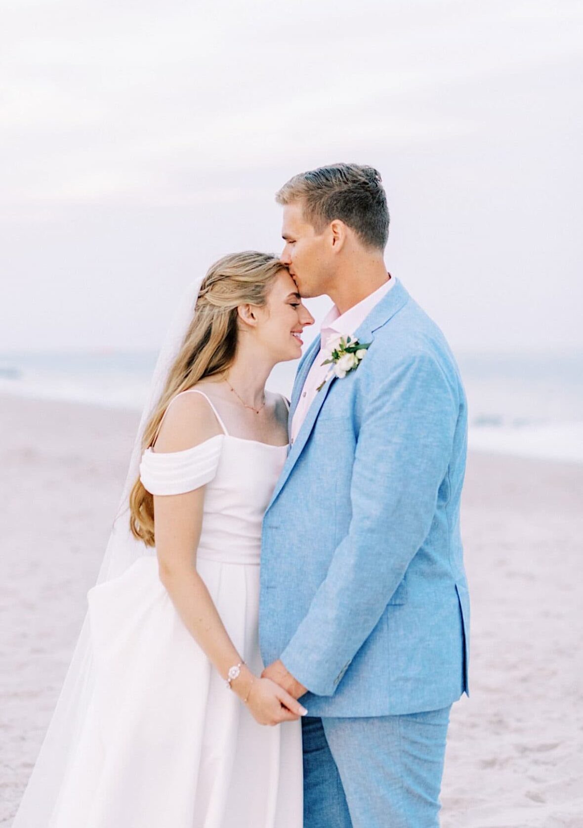 A bride and groom stand on a sandy beach at their Montreal Beach Resort wedding, embracing and smiling at each other in formal attire, with the ocean visible in the background.