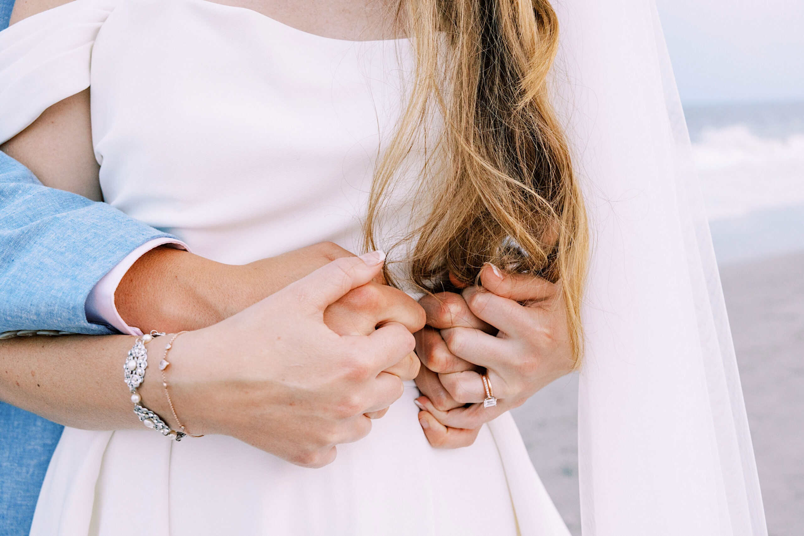 A couple stands close together on the beach at a Montreal Beach Resort wedding, holding hands. The person in white wears a bracelet and ring, while the person in blue has their arms around them.