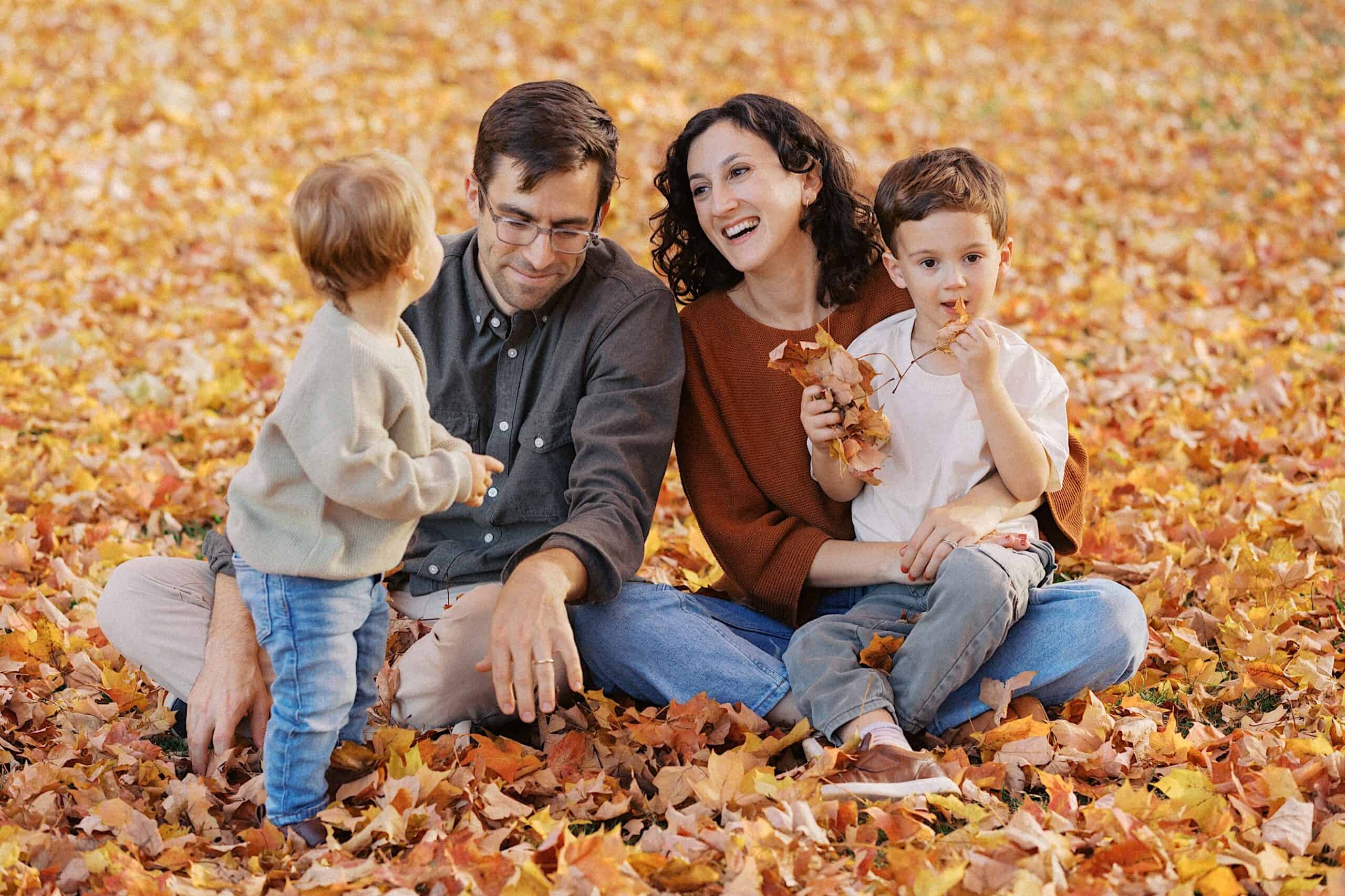 A family of four sits on the ground covered in autumn leaves. Two adults and two children are interacting and smiling among the fallen leaves.