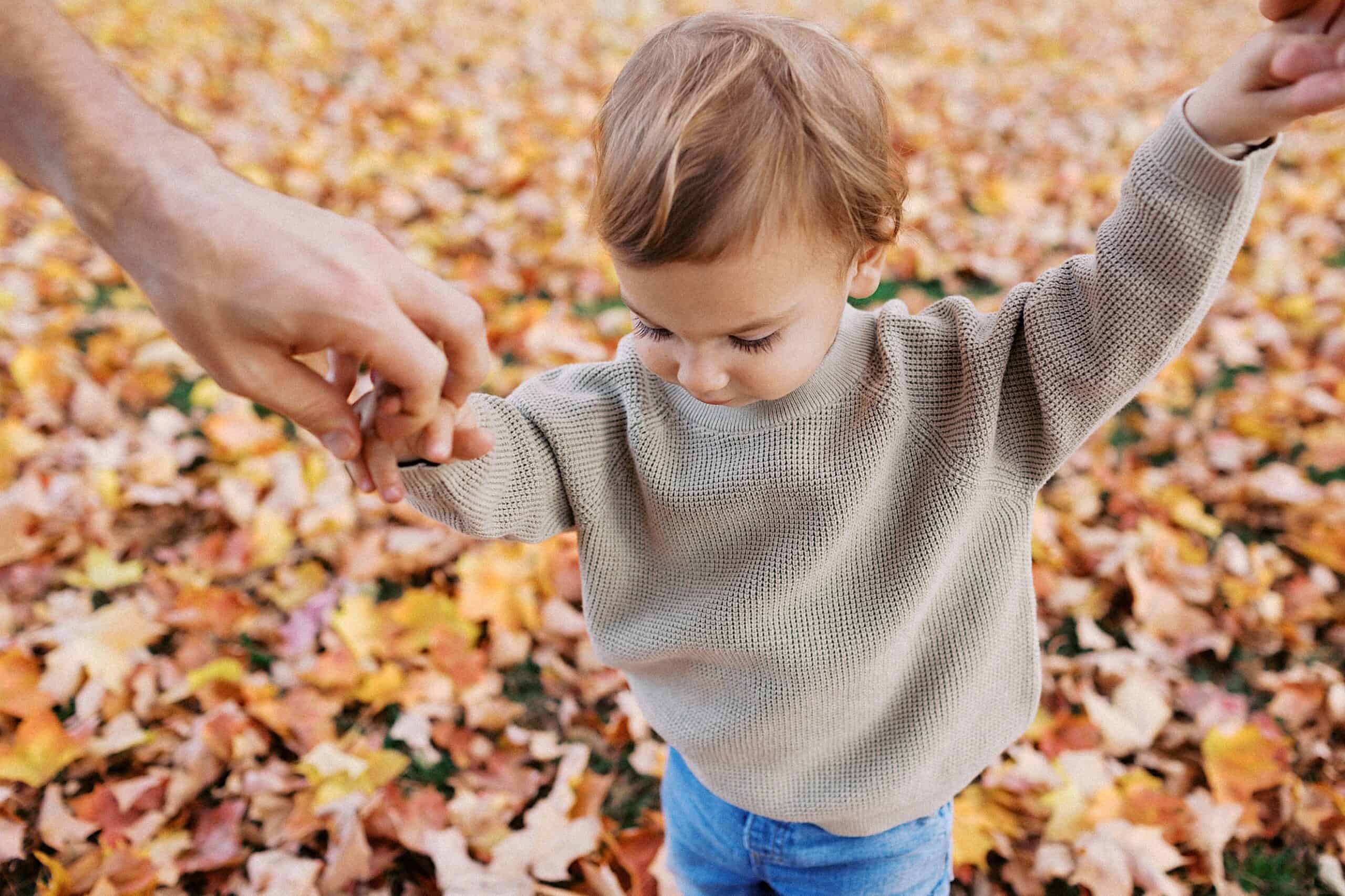 A young child in a beige sweater holds an adult's hand while walking through fallen autumn leaves.