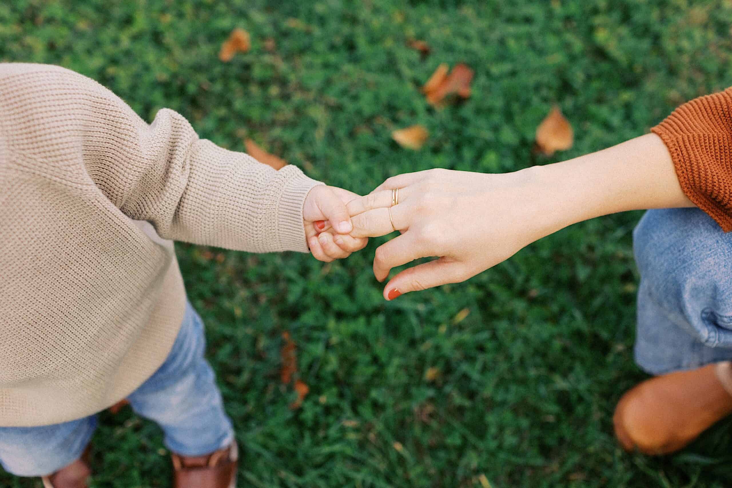 A child holding an adult's hand while standing on grass, both wearing sweaters and jeans.