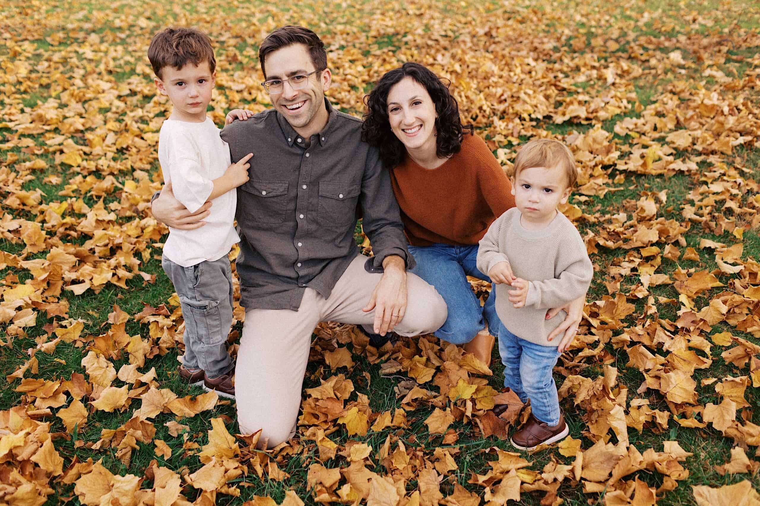 A family of four poses outdoors on grass covered with autumn leaves. An adult man kneels beside an adult woman, with two young children standing close to them.