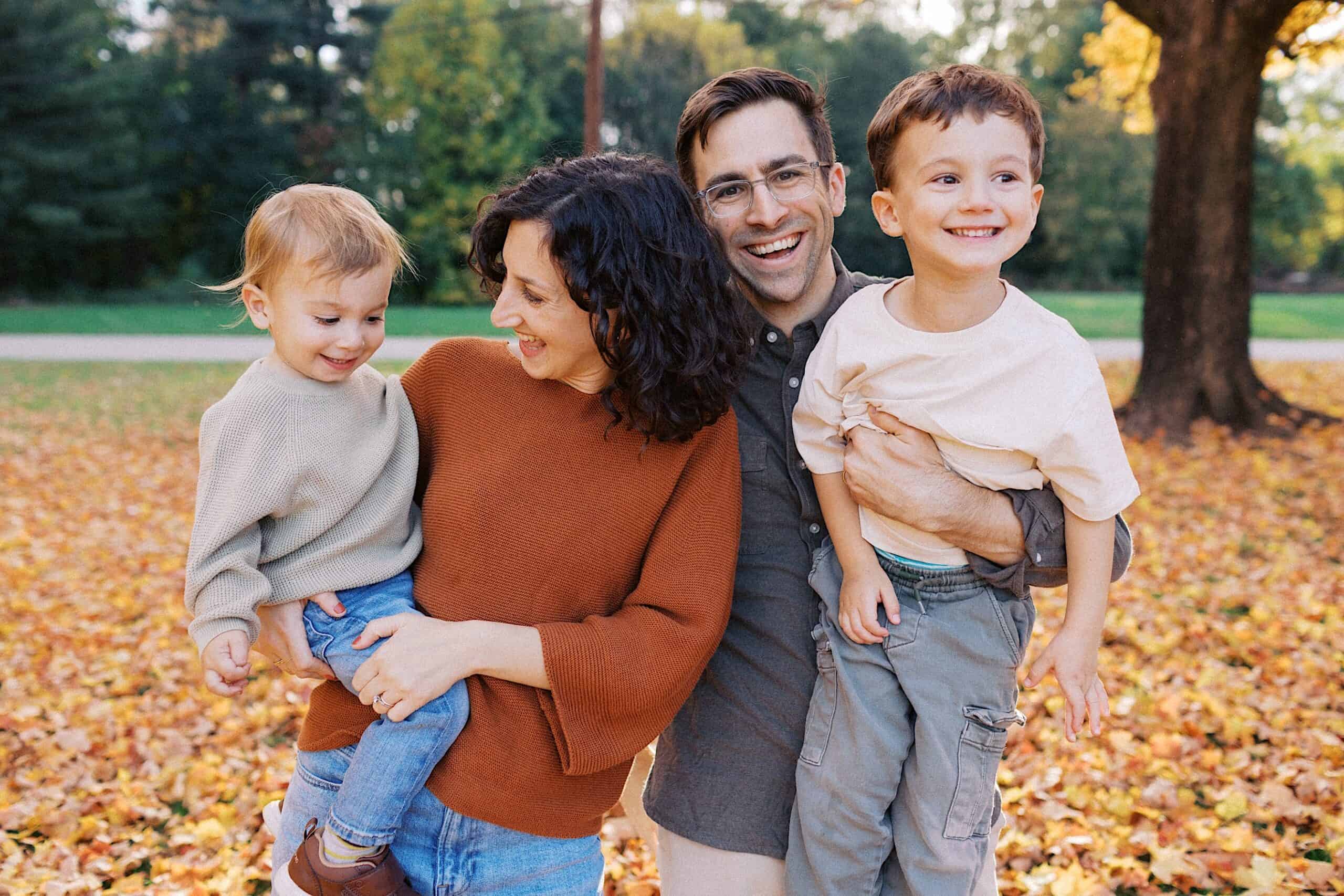 A family of four stands outdoors on fallen autumn leaves, smiling. Two adults hold two young children, with trees and greenery in the background.