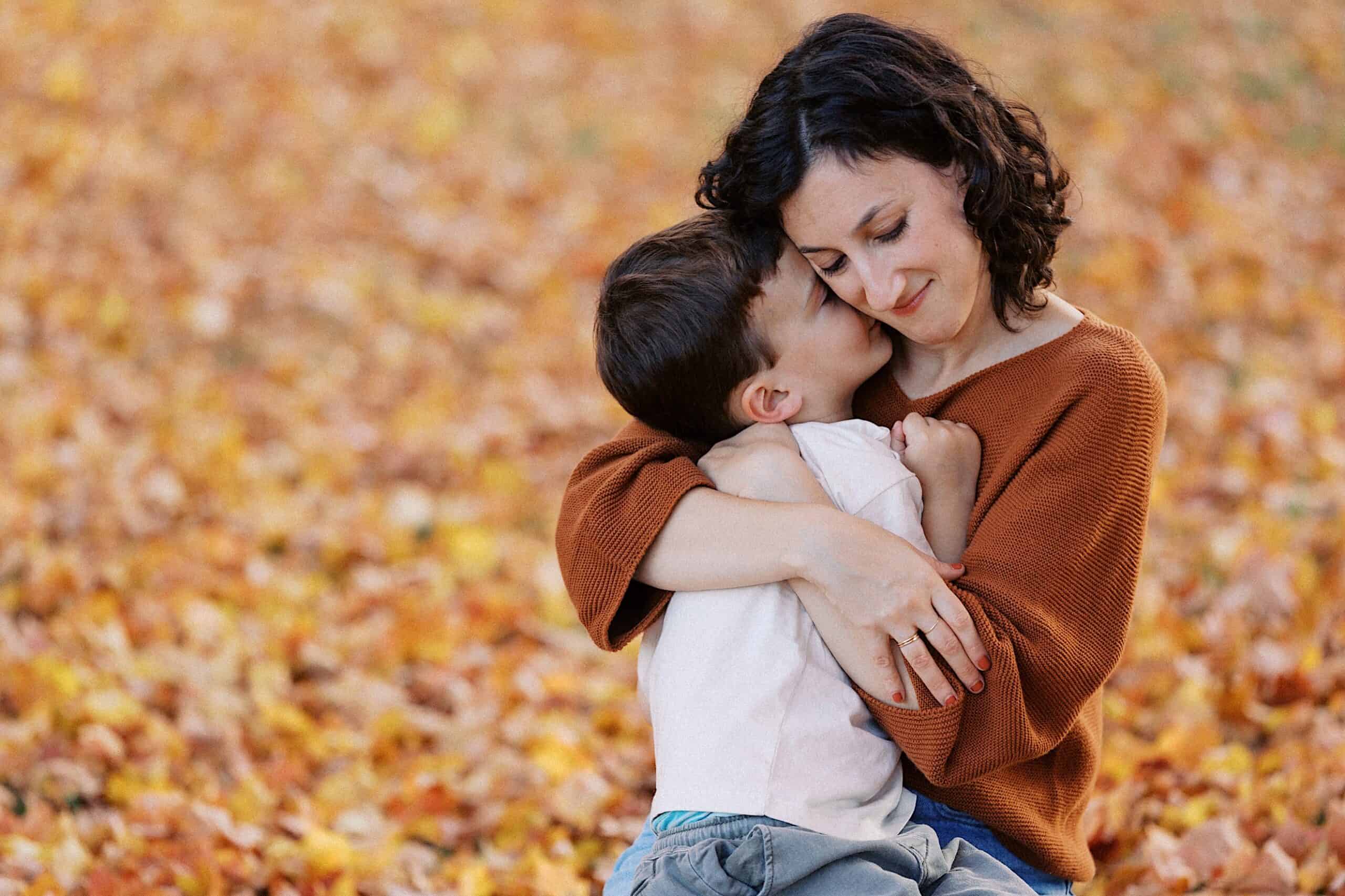Woman in a brown sweater hugs a young boy on the ground covered with autumn leaves. The boy’s face is pressed against her cheek and she appears calm and content.