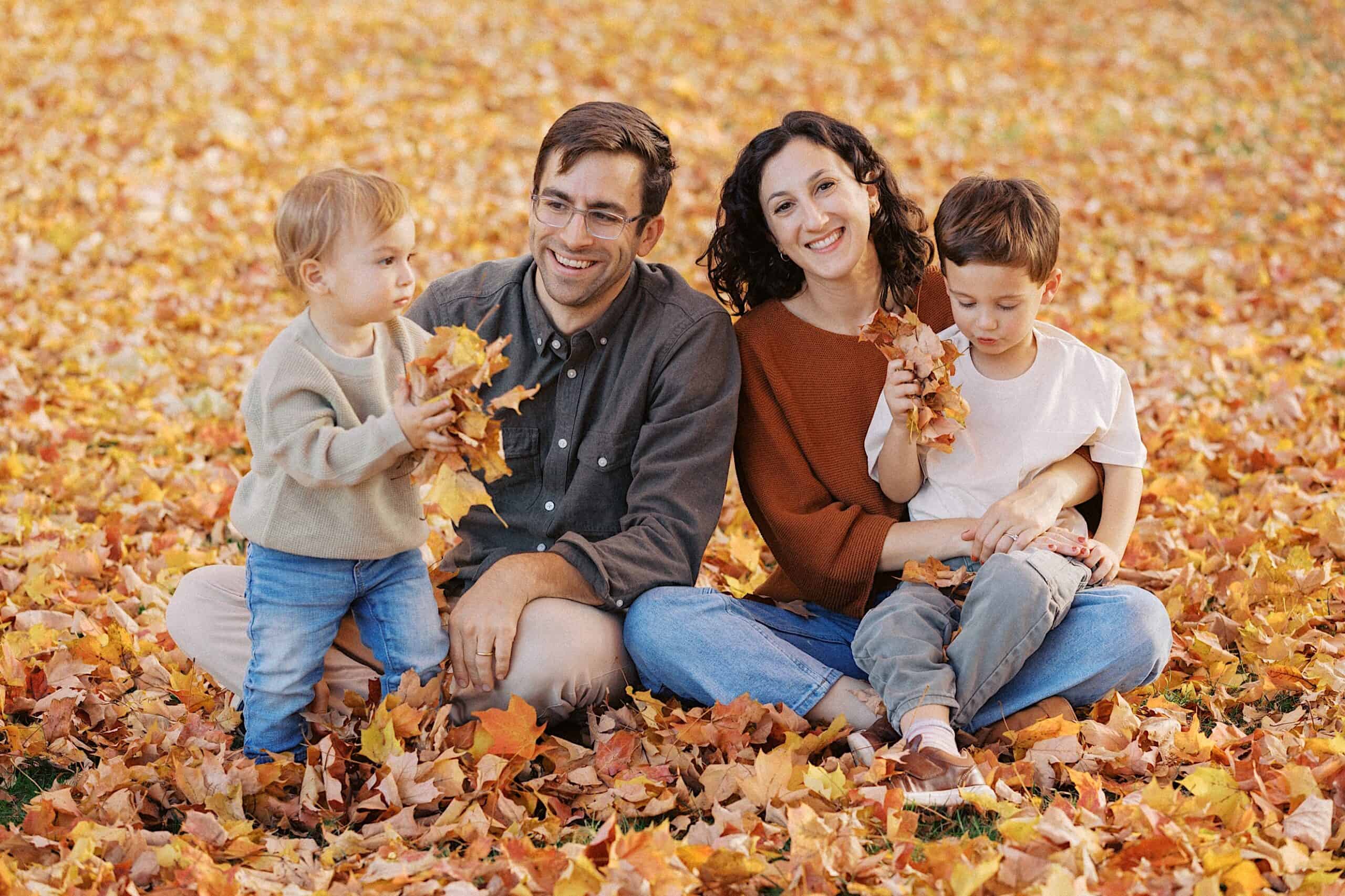 A family of four sits on the ground covered in autumn leaves, with two young children holding handfuls of leaves and two adults smiling at the camera.
