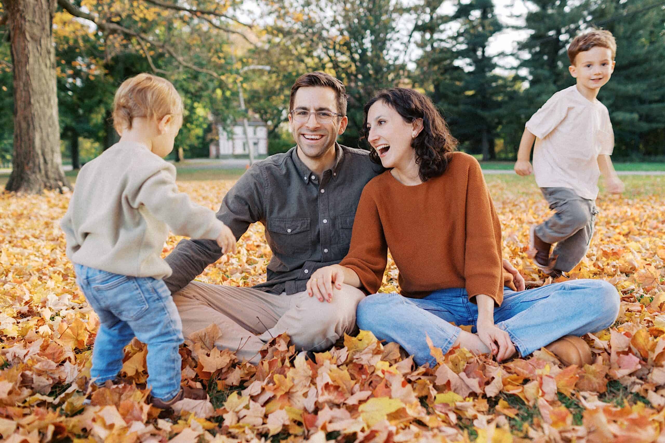 A couple sits on autumn leaves in a park, smiling and holding hands, while two young children play nearby.