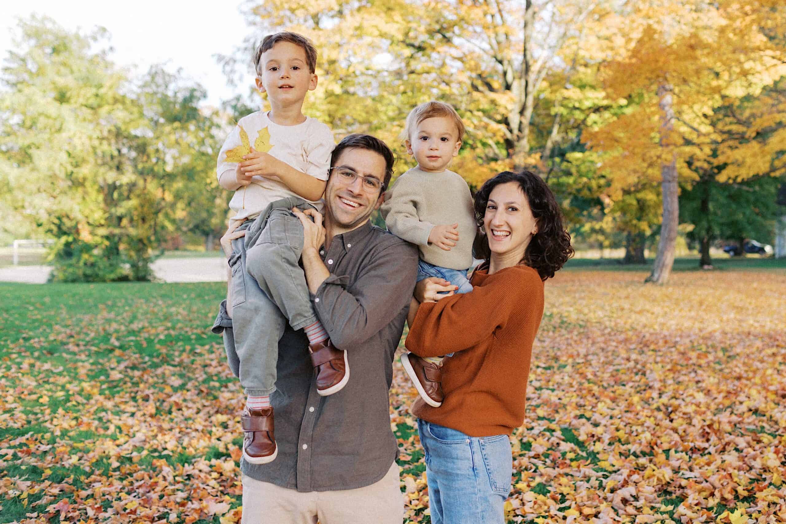 Two adults and two children pose together in a park with autumn leaves on the ground and colorful trees in the background.