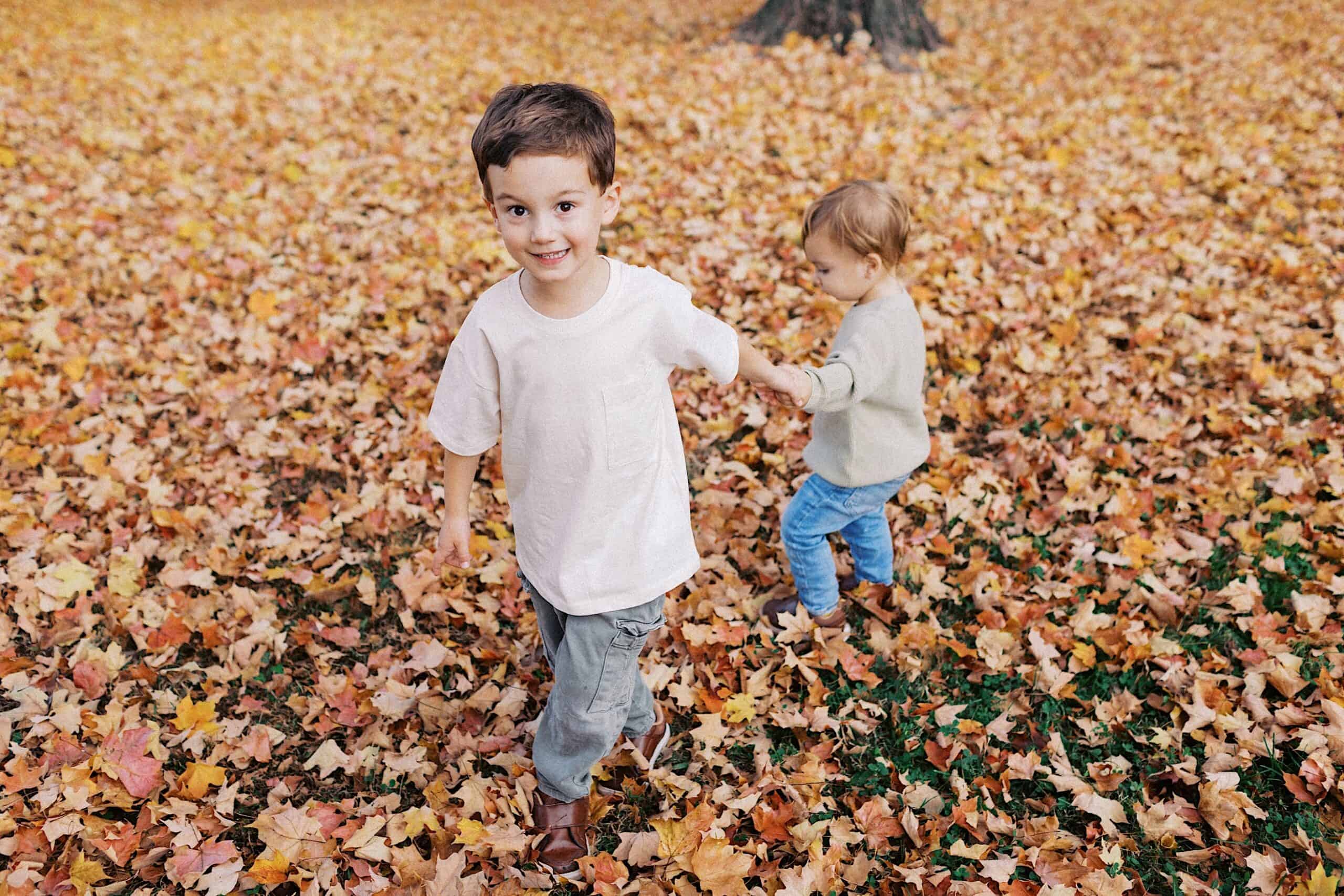 Two young children walk through fallen autumn leaves; the older child smiles at the camera while holding the younger child’s hand.