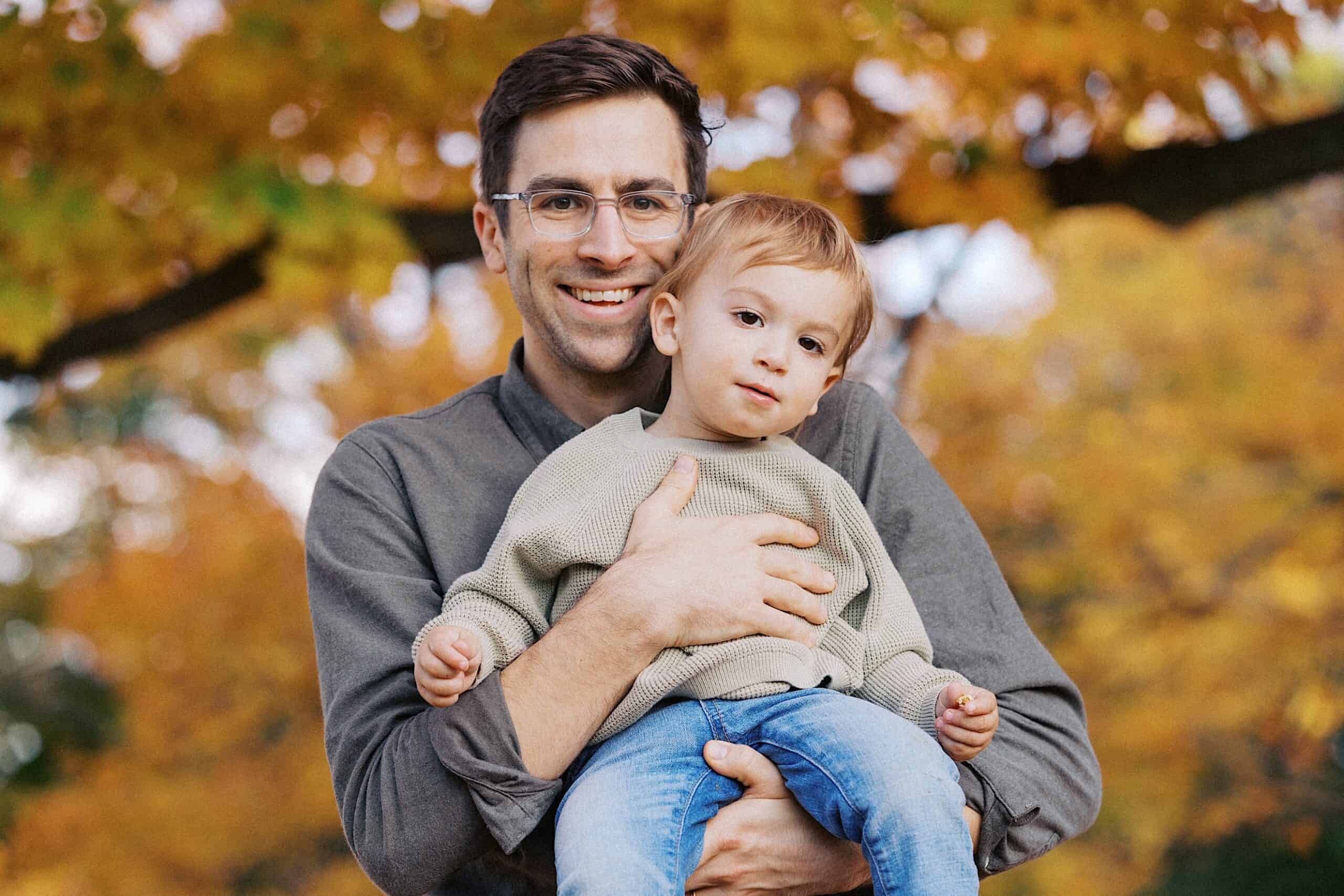 An adult holding a small child outdoors, both facing the camera, with autumn-colored trees in the background.