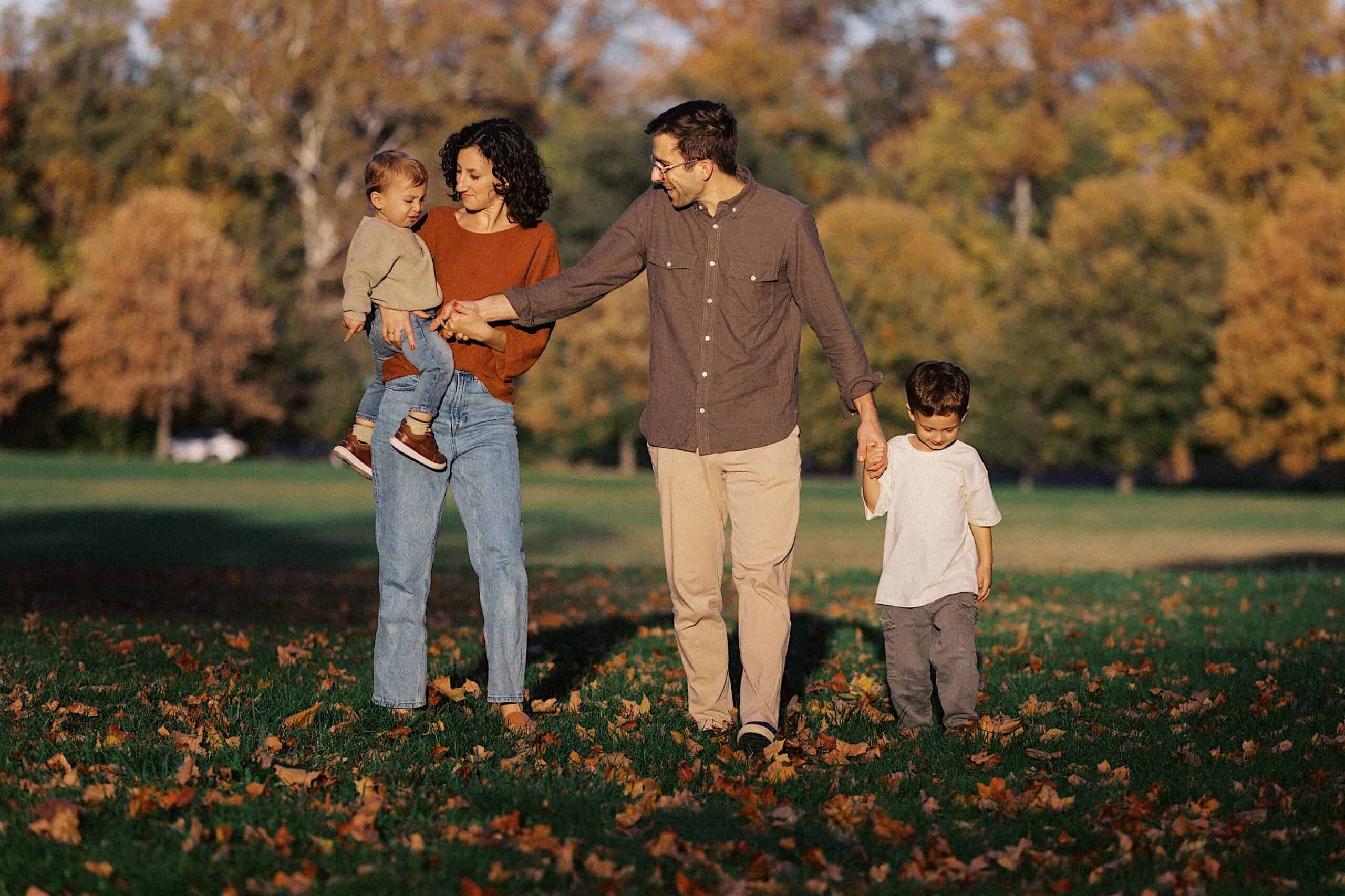 A family of four walks together on a grassy field with autumn leaves, surrounded by trees with fall foliage.