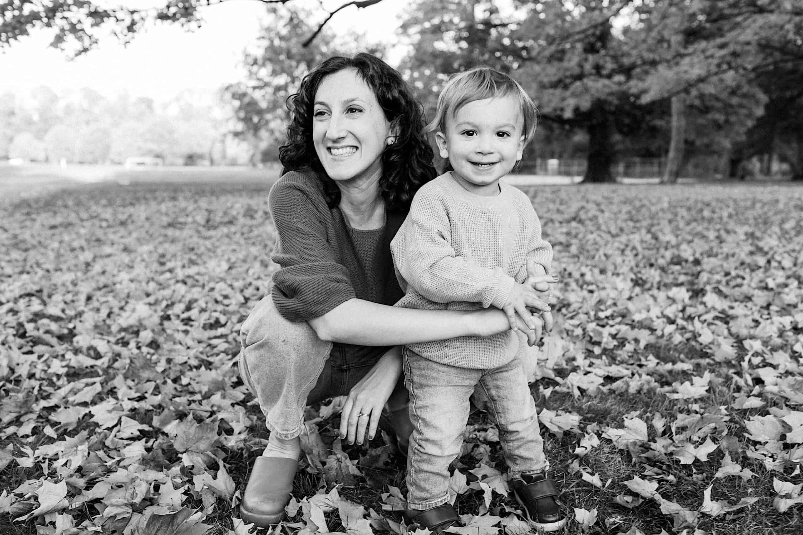 A woman and a young child smile while posing together outdoors on a ground covered with fallen leaves.