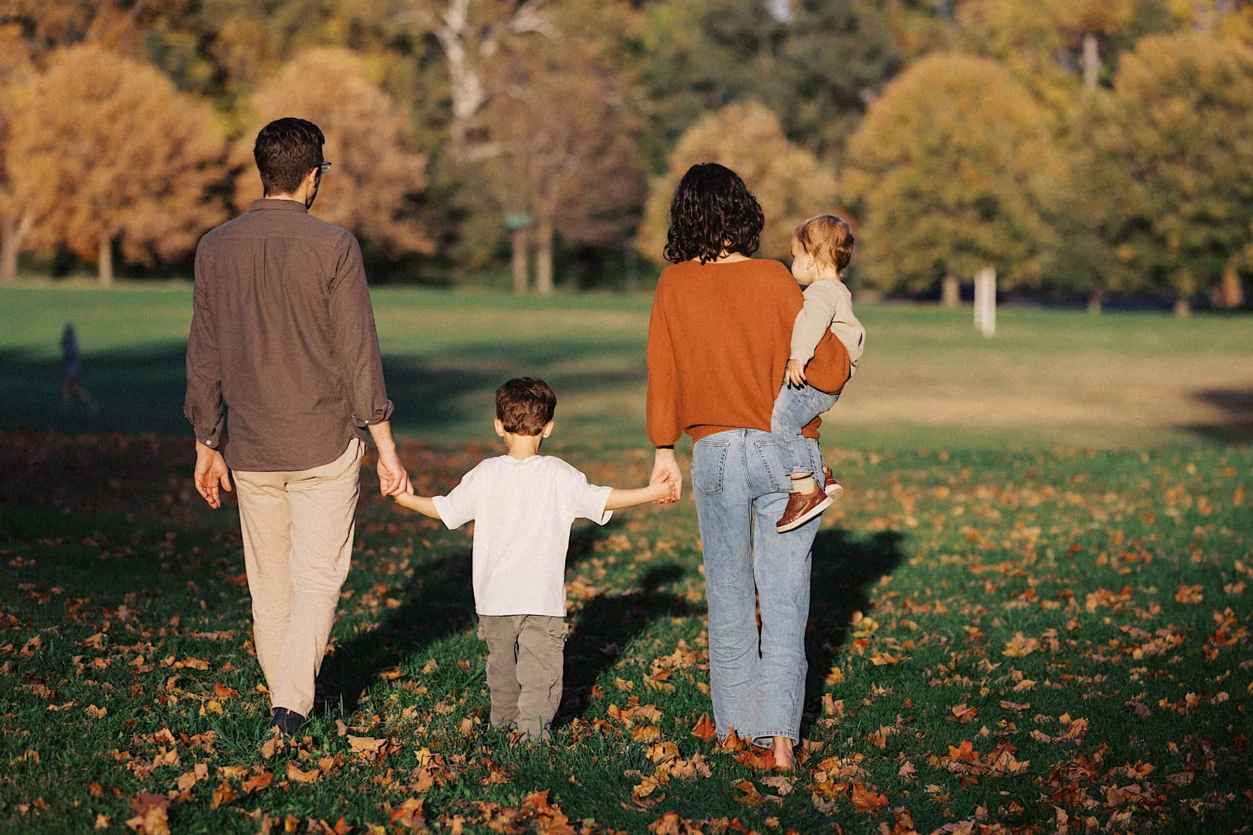 A family of four walks away on a grassy field with autumn leaves, two adults holding hands with a child, one adult carrying a toddler. Trees with fall foliage are in the background.