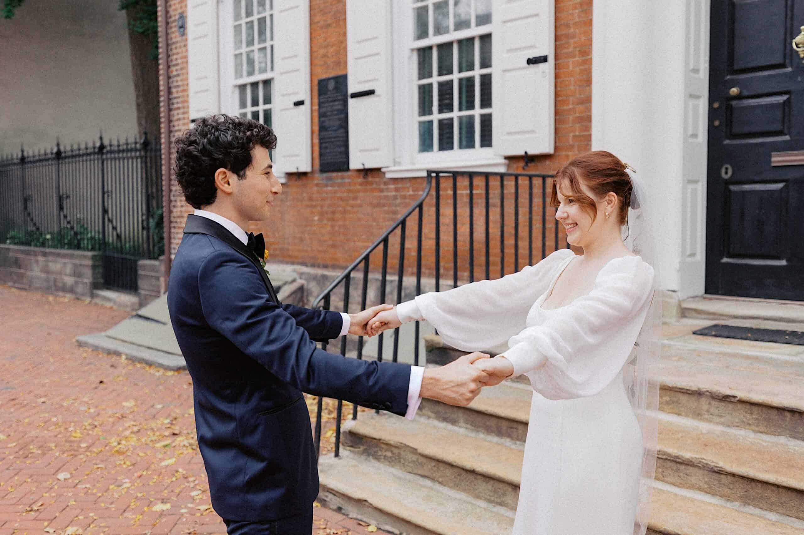 A bride and groom stand outside a brick building, holding hands and smiling at each other on a paved path with scattered leaves.