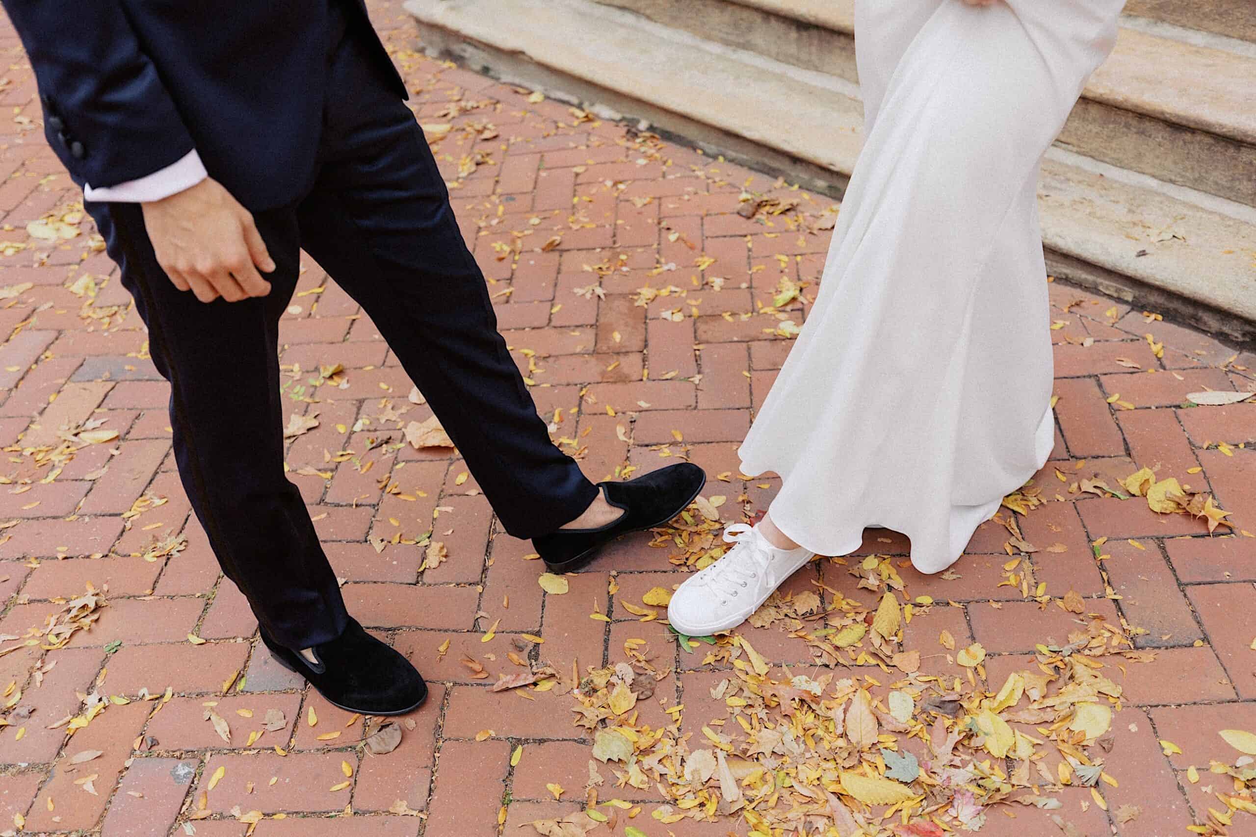 Two people stand on a brick path with fallen leaves; one wears a dark suit and loafers, the other wears a white dress and white sneakers.