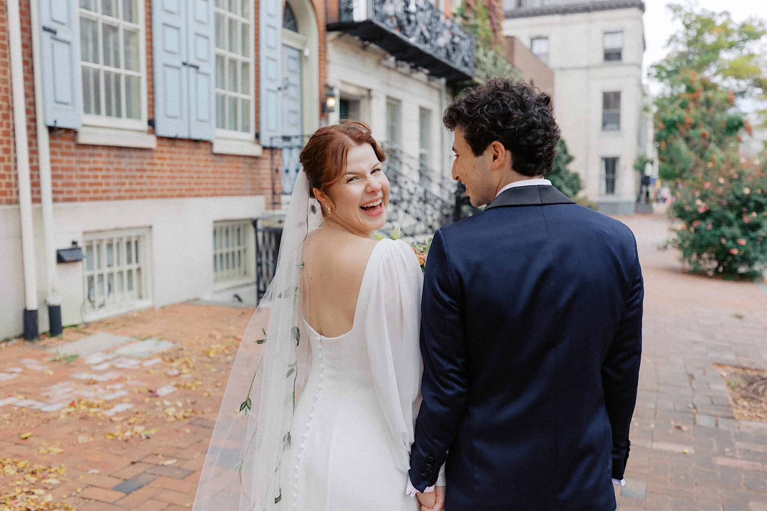 A bride and groom stand outdoors holding hands, smiling and looking back at the camera on a brick walkway beside historic buildings.
