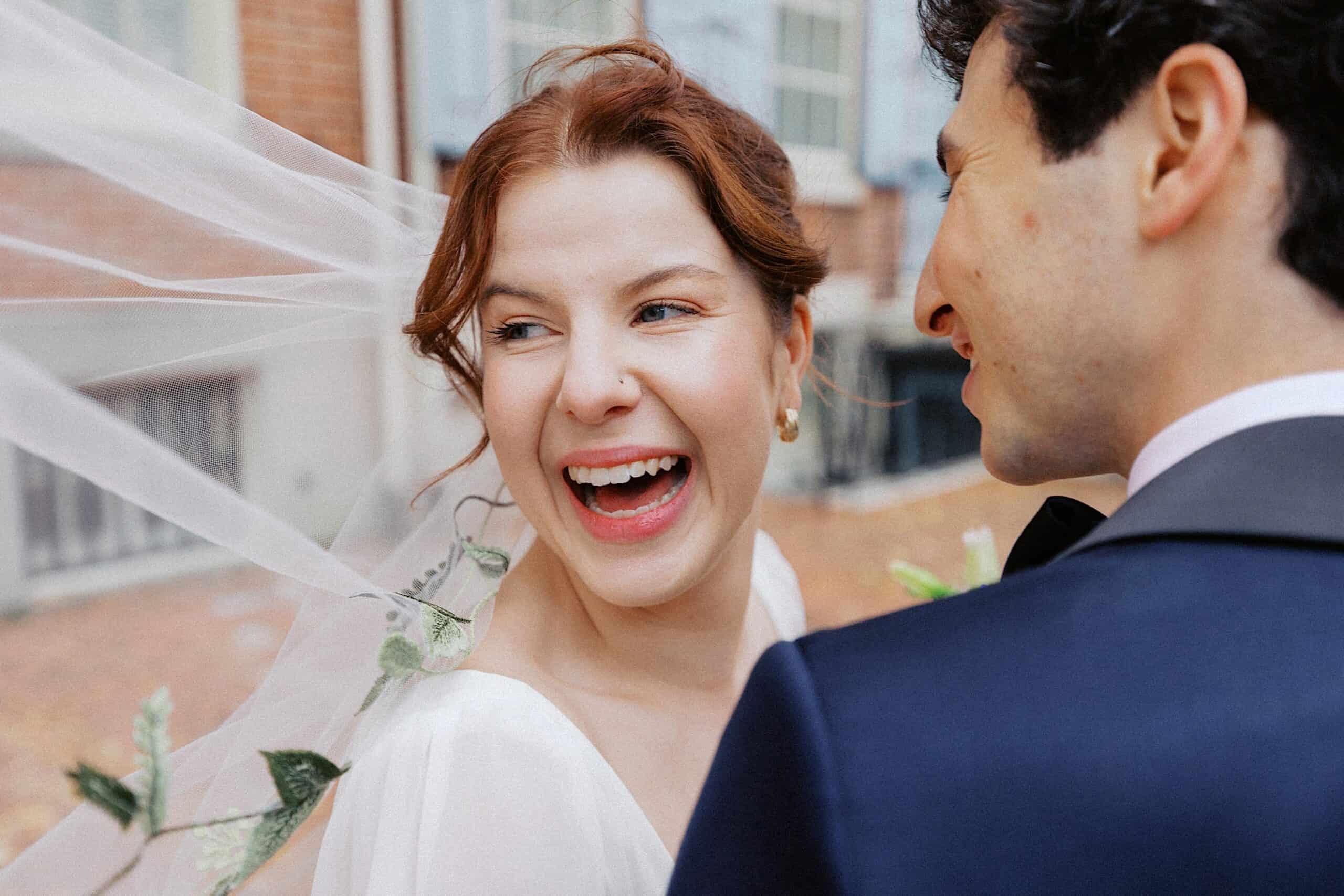 A bride in a white dress smiles joyfully while standing next to a groom in a dark suit outdoors. Her veil is flowing in the breeze.