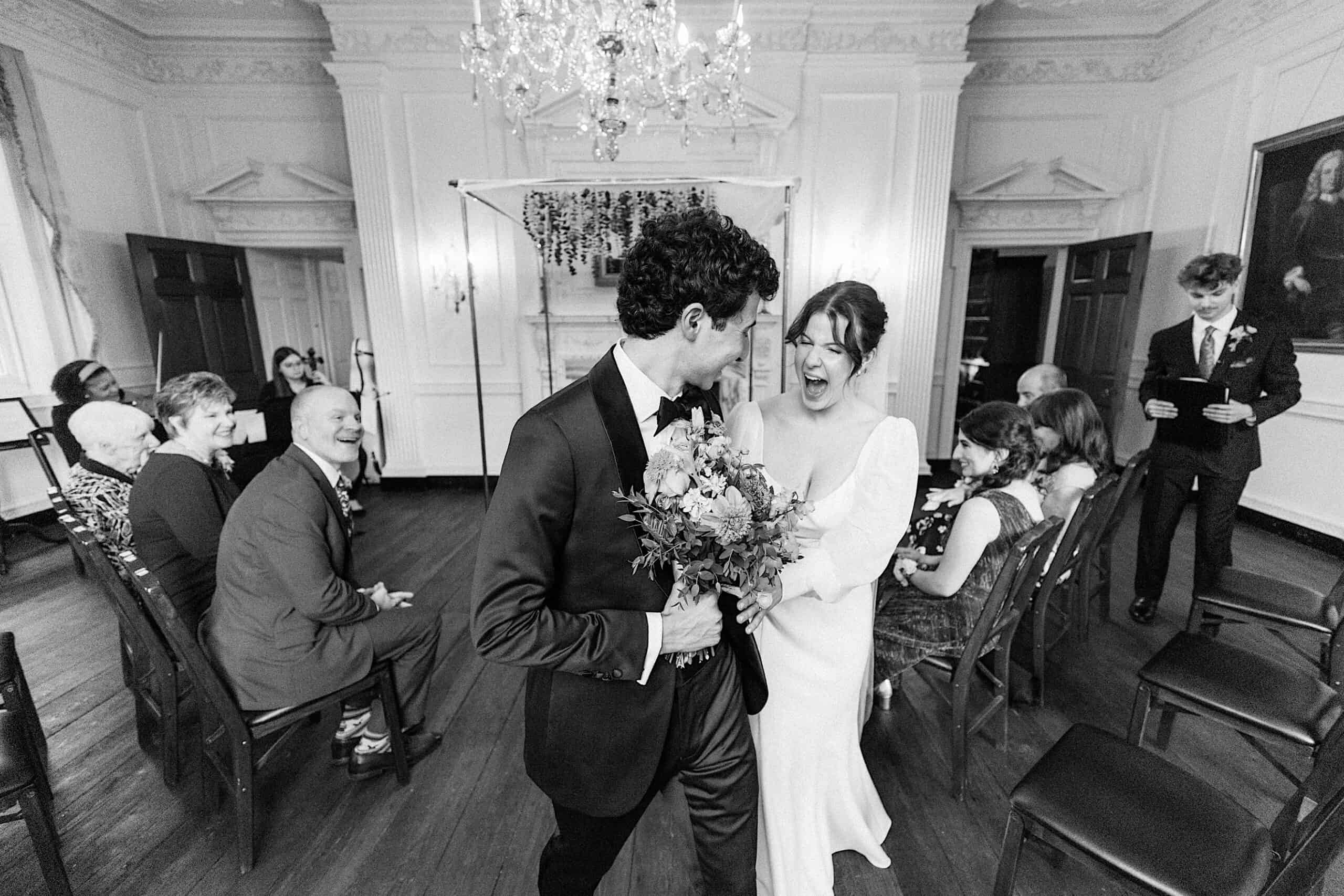 A bride and groom walk down the aisle smiling, holding a bouquet, as wedding guests seated on both sides watch in a decorated indoor venue.