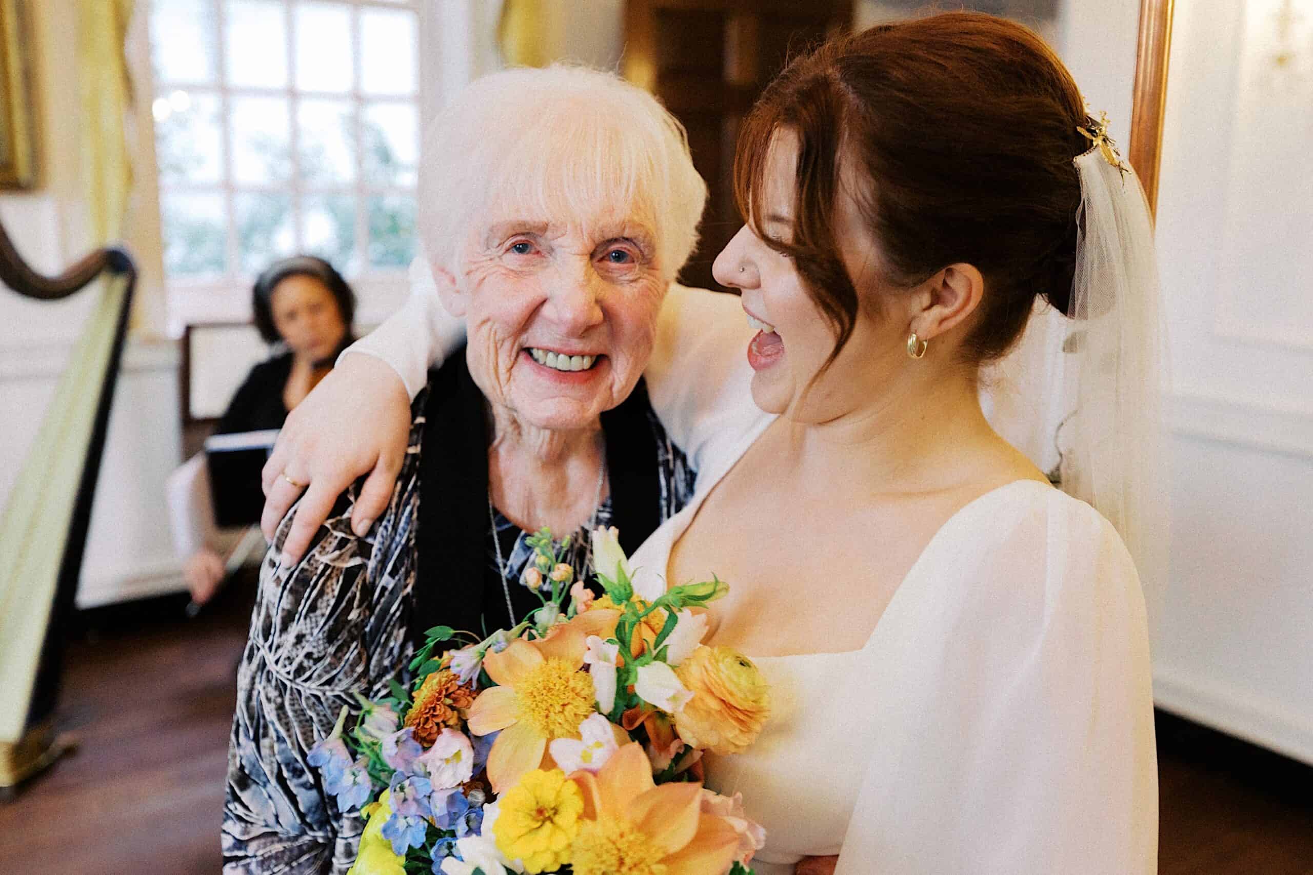 An elderly woman and a bride smile and pose together indoors; the bride holds a colorful bouquet and a harpist is seen in the background.