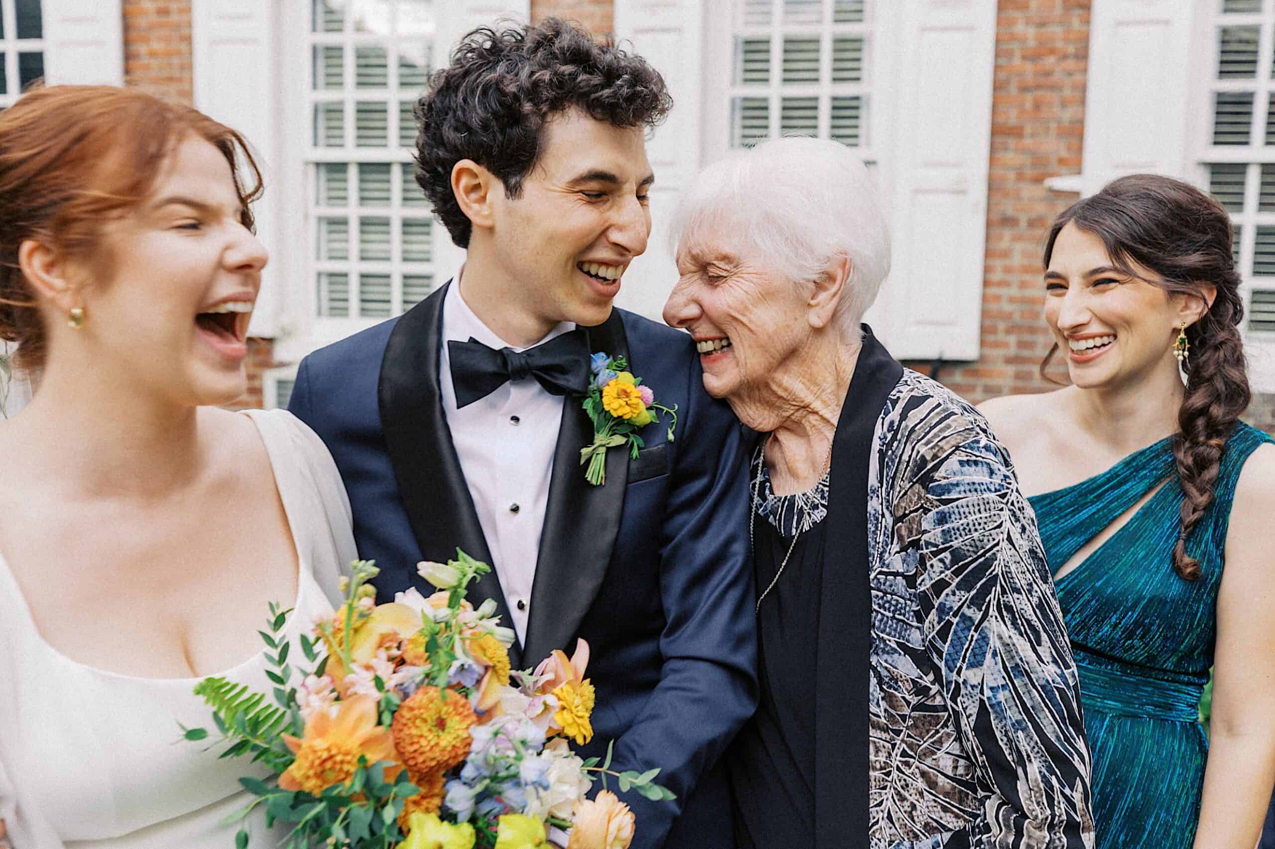 Four people, including a groom in a tuxedo holding a bouquet, smile and laugh together outdoors in front of a brick building with white windows.