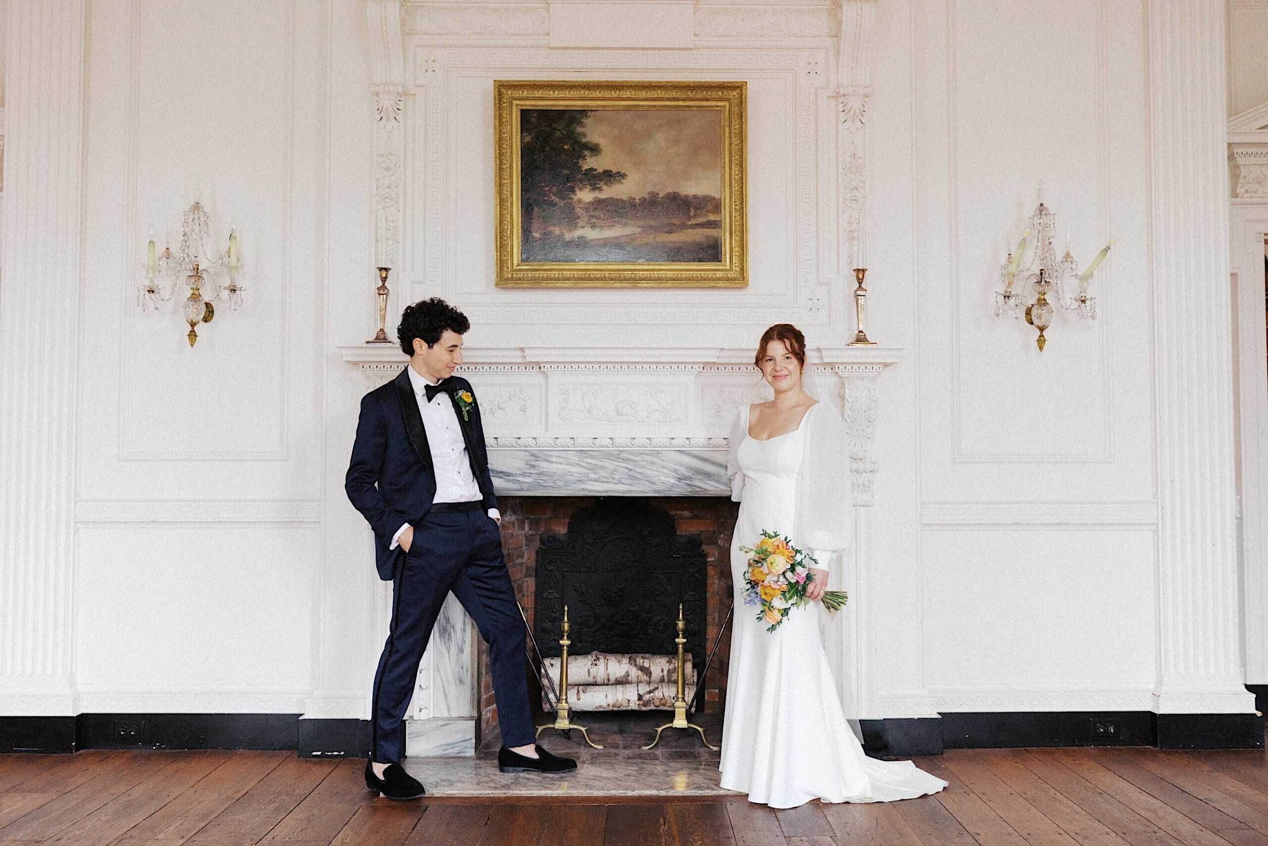 A bride in a white dress holding a bouquet stands next to a groom in a dark suit in front of a fireplace in an elegant room.