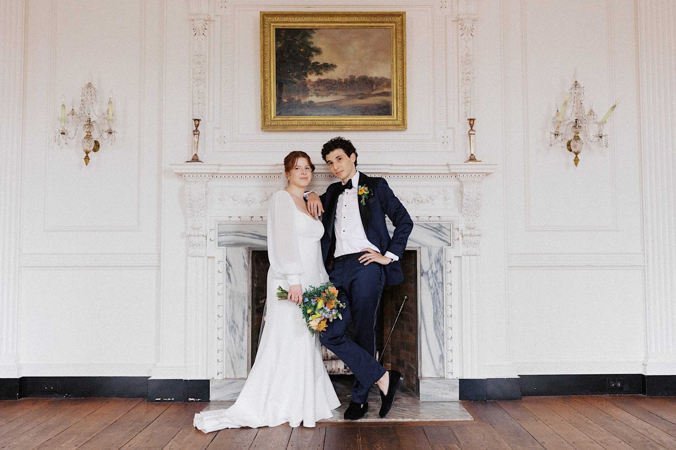 A bride in a white dress holds a bouquet, standing beside a person in a dark suit; both pose in front of a fireplace in an elegant, white-walled room with a framed painting above.