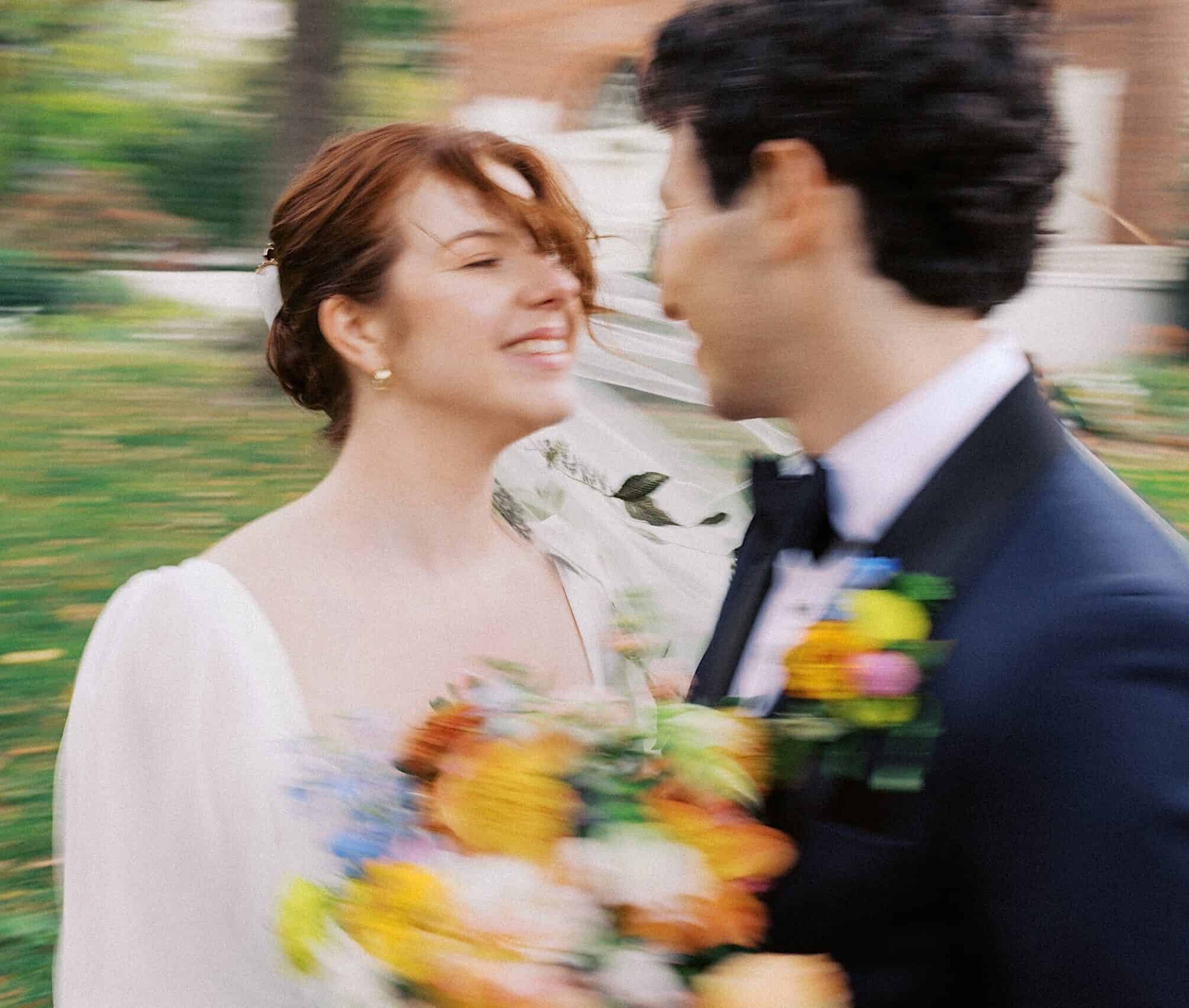 A bride and groom stand close together outdoors, smiling and holding a colorful bouquet, with a blurred background of greenery and a building.