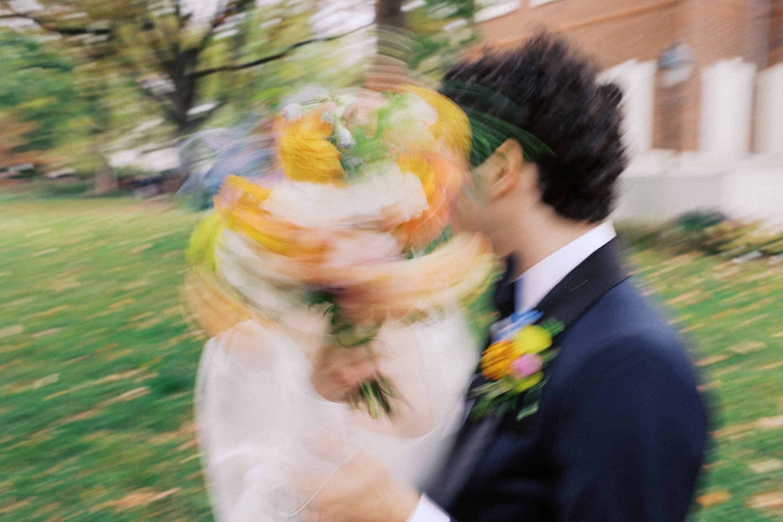 A bride and groom stand outside, faces obscured by motion blur as the bride holds a bouquet up, with greenery and a building in the background.