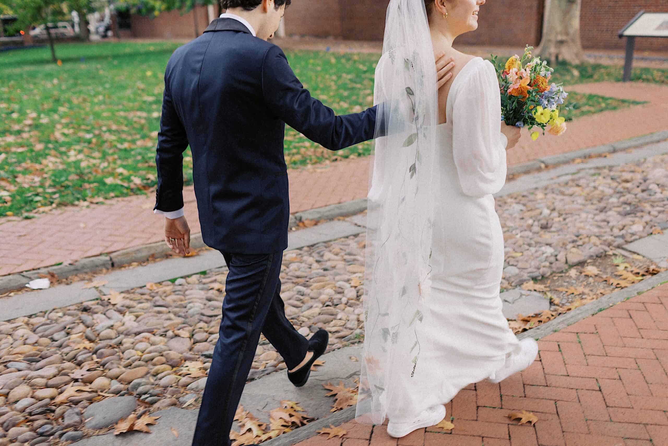 A bride in a white dress and veil walks with a groom in a dark suit on a brick path outdoors, holding a bouquet of flowers.