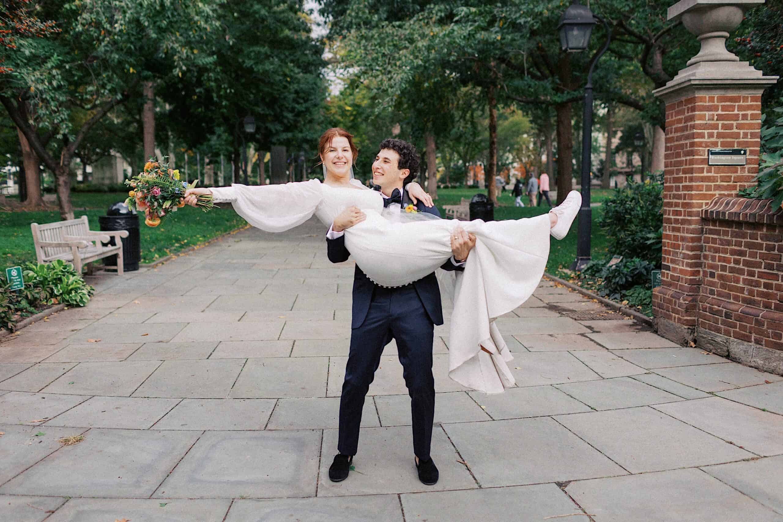 A person in a suit lifts a person in a white dress, holding a bouquet, on a stone path in a park with trees and brick columns.