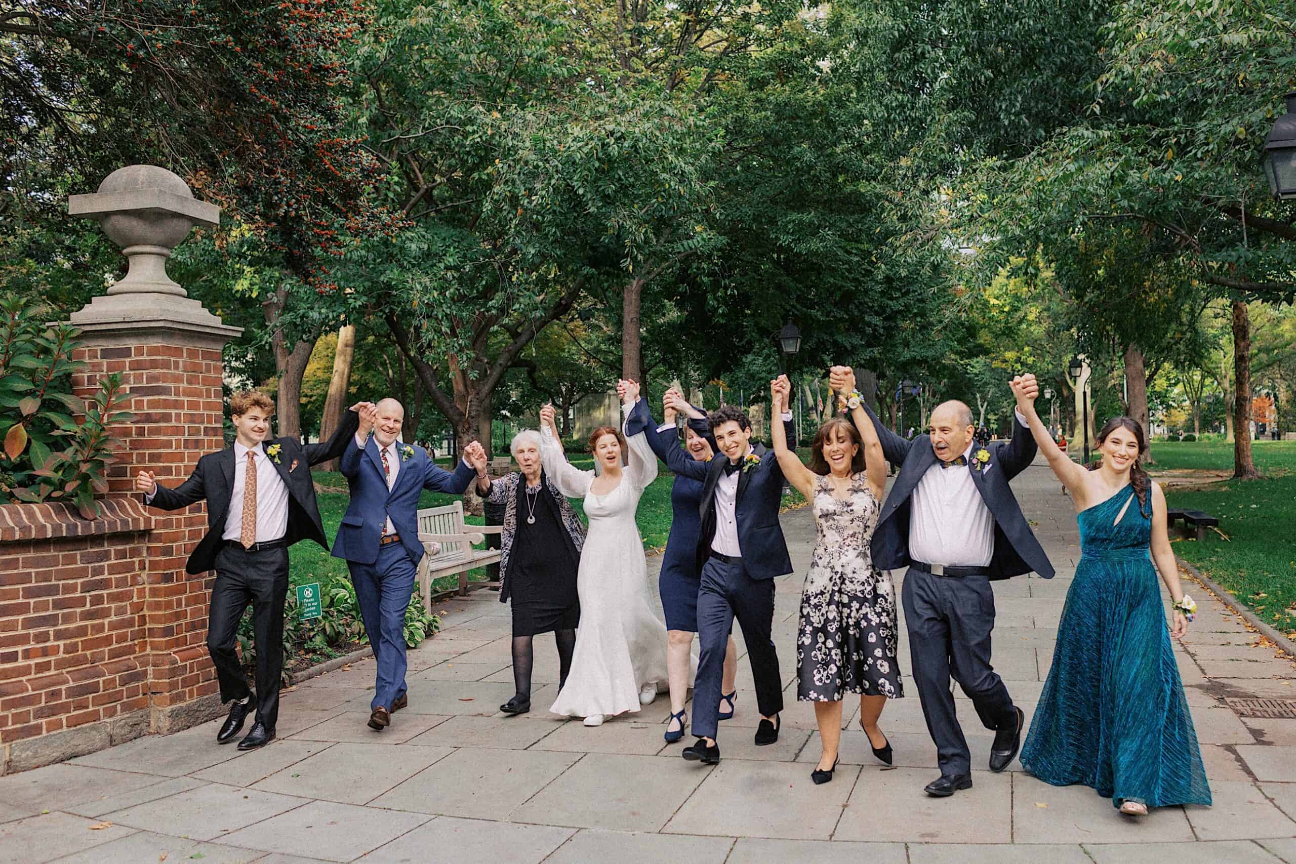 A group of formally dressed people, including a bride and groom, walk outside on a paved path, holding hands and raising their arms in celebration.