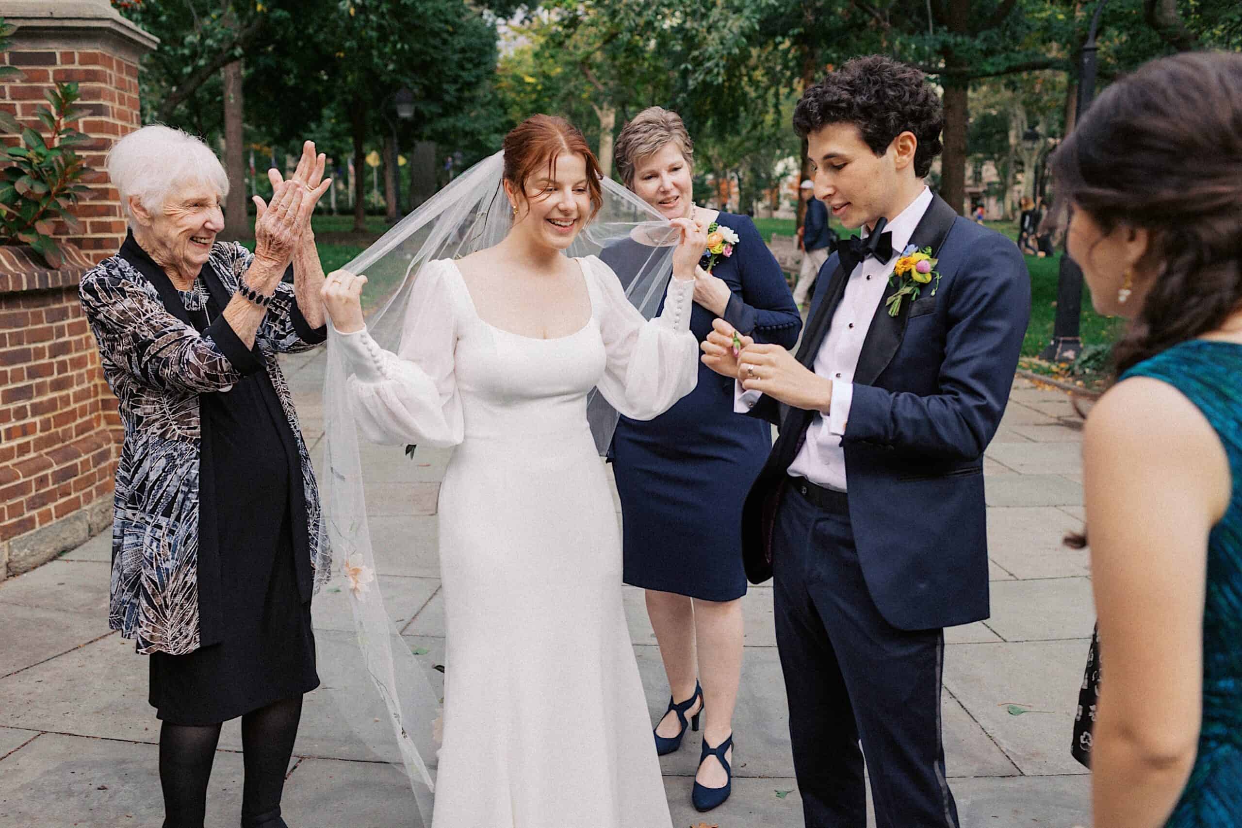 A bride in a white dress stands with a groom in a suit outdoors, surrounded by three guests; one older woman is clapping and smiling.